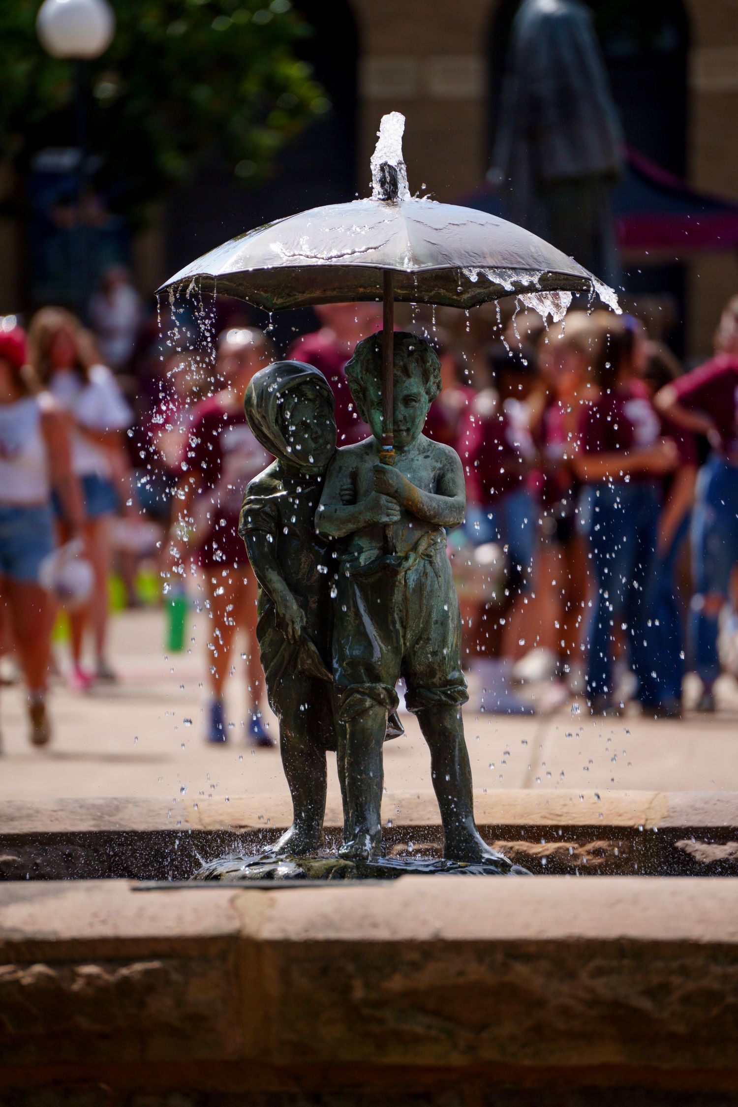 Bronze fountain sculpture of two children sharing an umbrella with water splashing, set in a busy public square.
