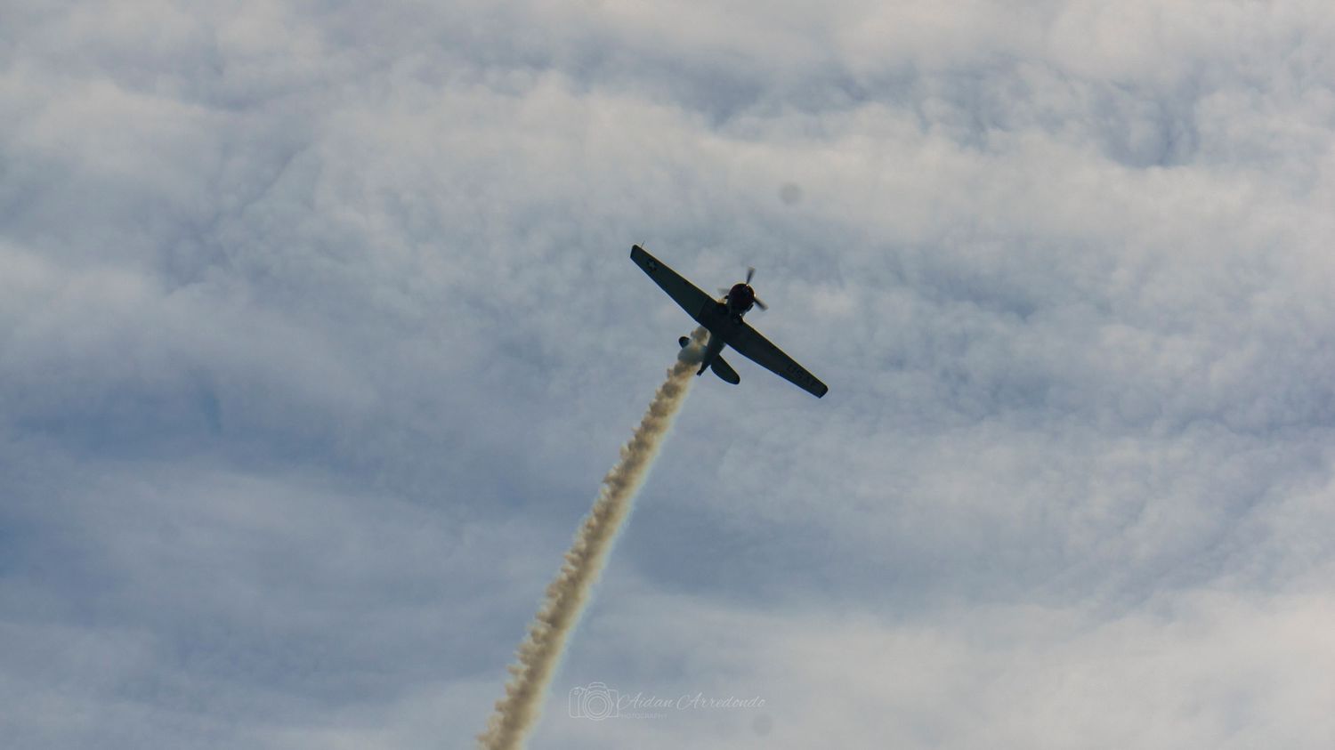 A small aircraft performs aerobatics leaving a smoke trail against a cloudy blue sky.