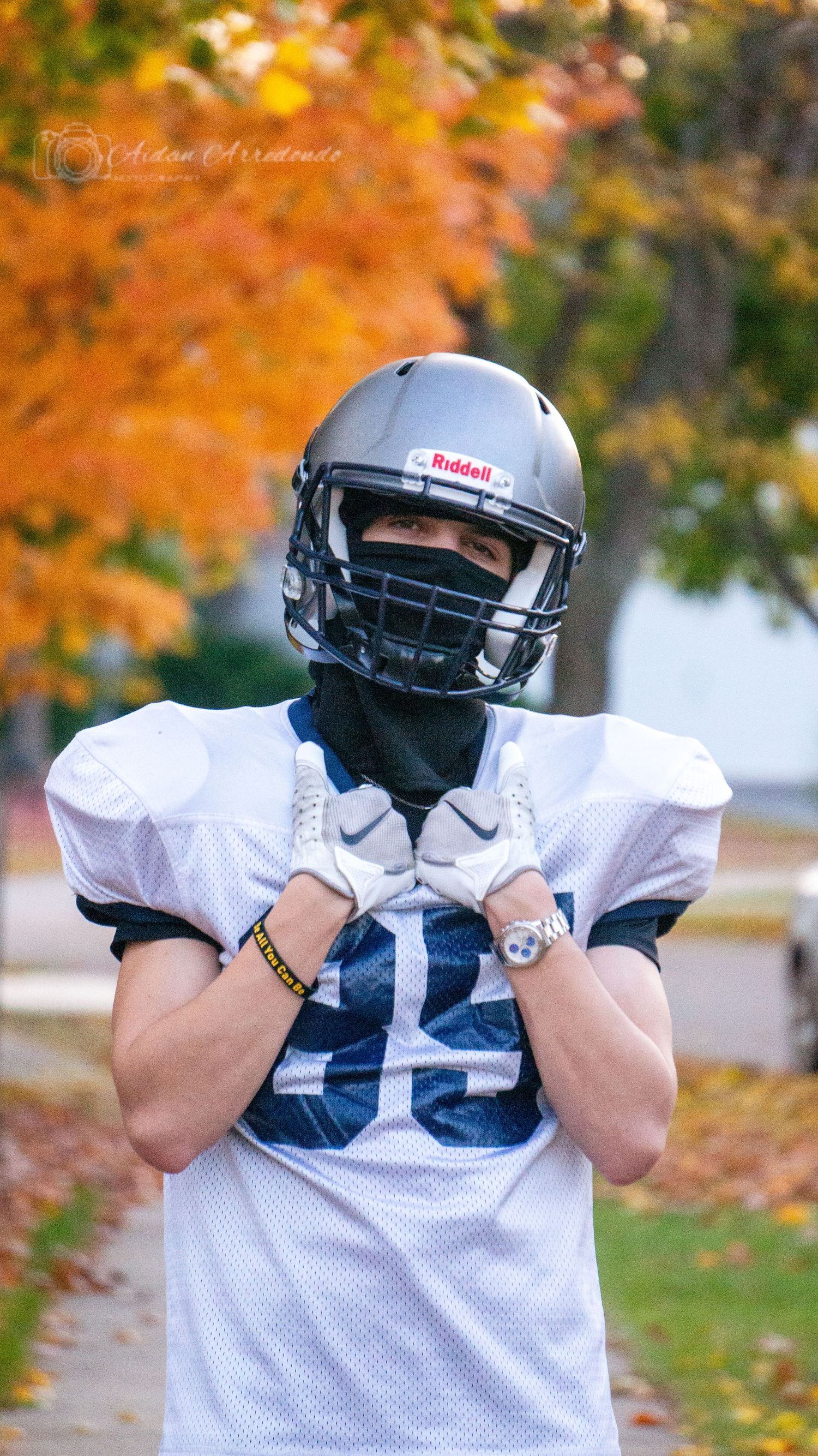 Football player in full uniform and Riddell helmet poses on a tree-lined autumn path.
