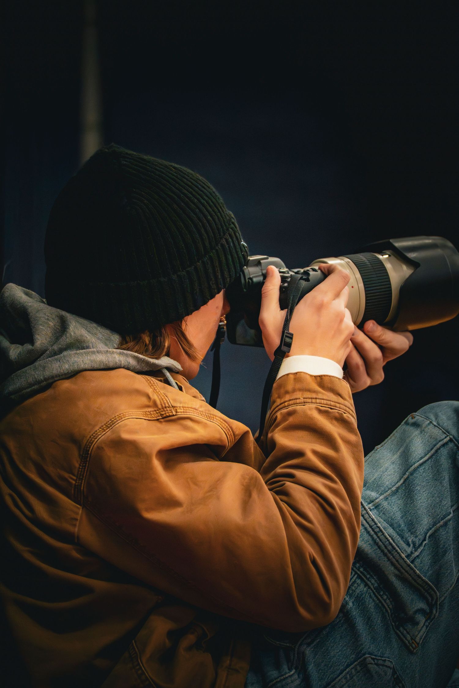 Photographer in brown leather jacket and black beanie captures shots with a telephoto lens camera in low light.