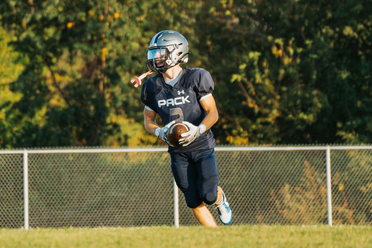 Youth football player in dark Pack uniform catches ball during outdoor game practice on sunny day.