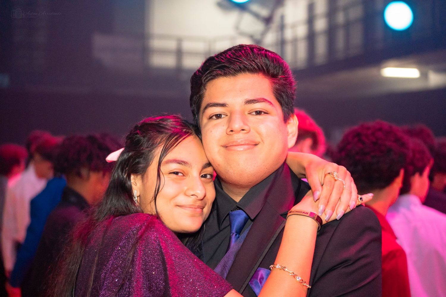 Two teens smiling and hugging at a school dance event under colorful lights.