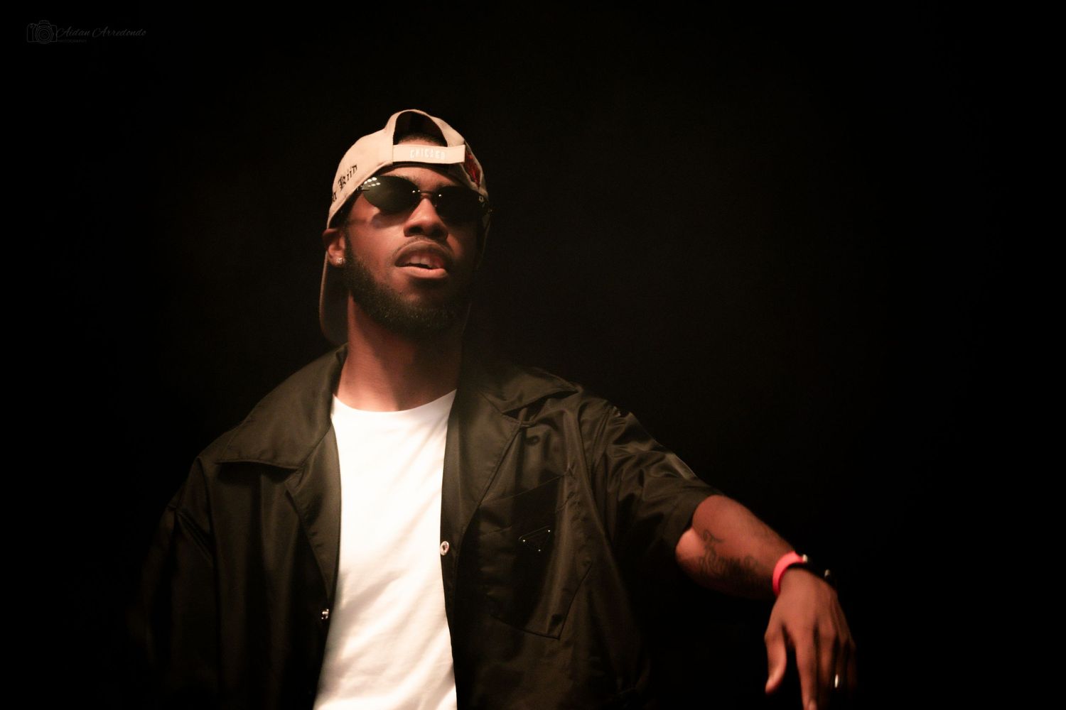 Young man in a dark jacket and backwards cap poses against a dark studio background.