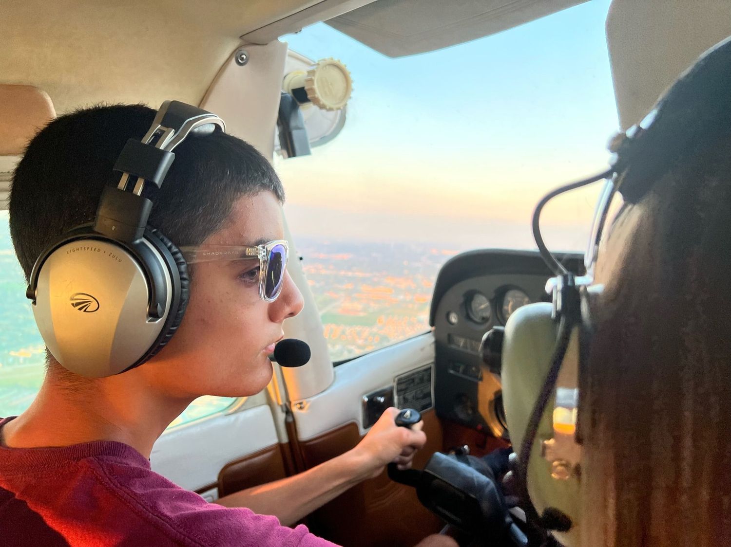Young boy wearing headphones learning to fly a small aircraft with an instructor at sunset.