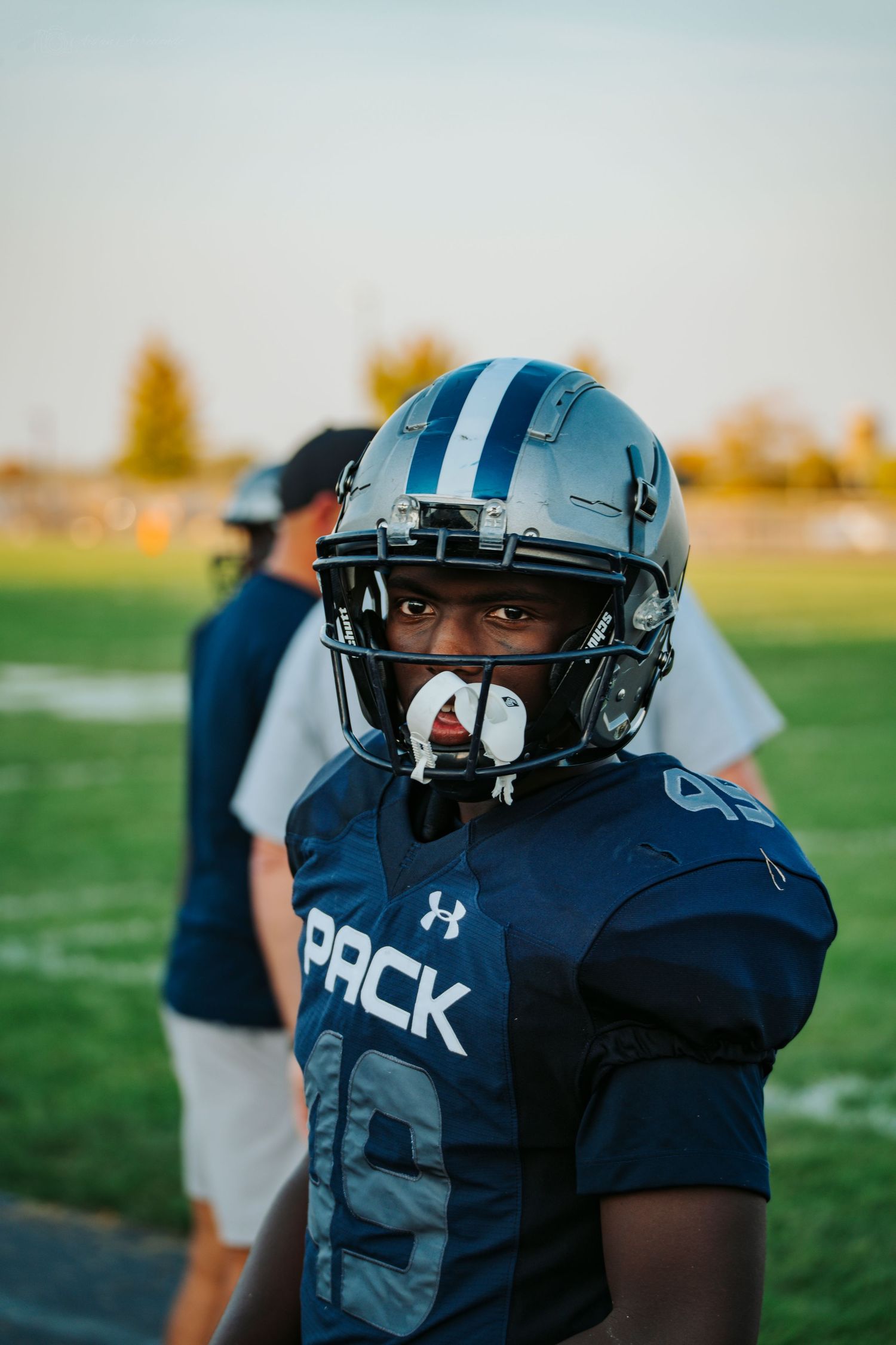 Young football player in navy Pack uniform and helmet stands on field at sunset during game.