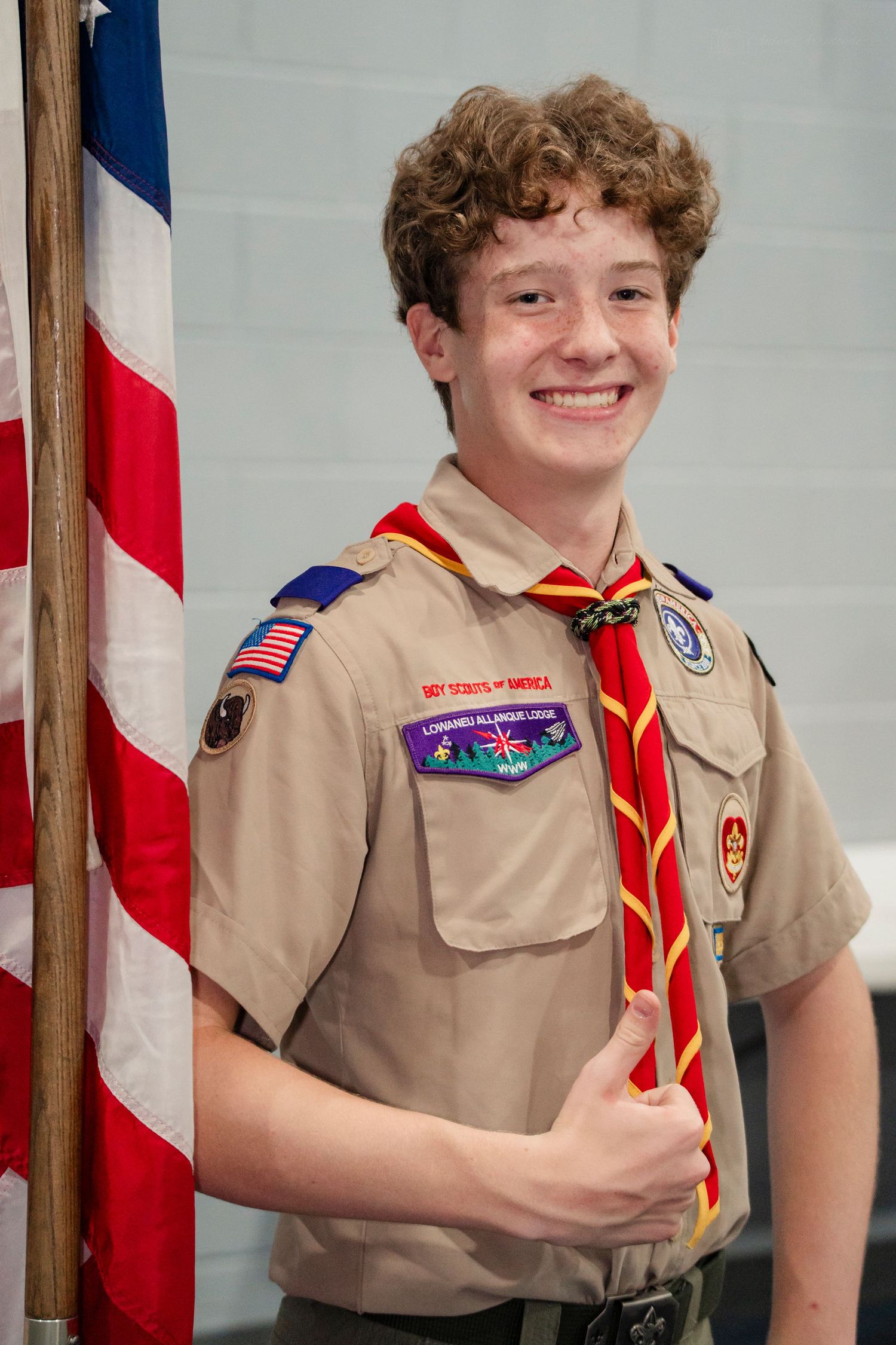 Smiling teenage Boy Scout in uniform holds an American flag giving a thumbs up.