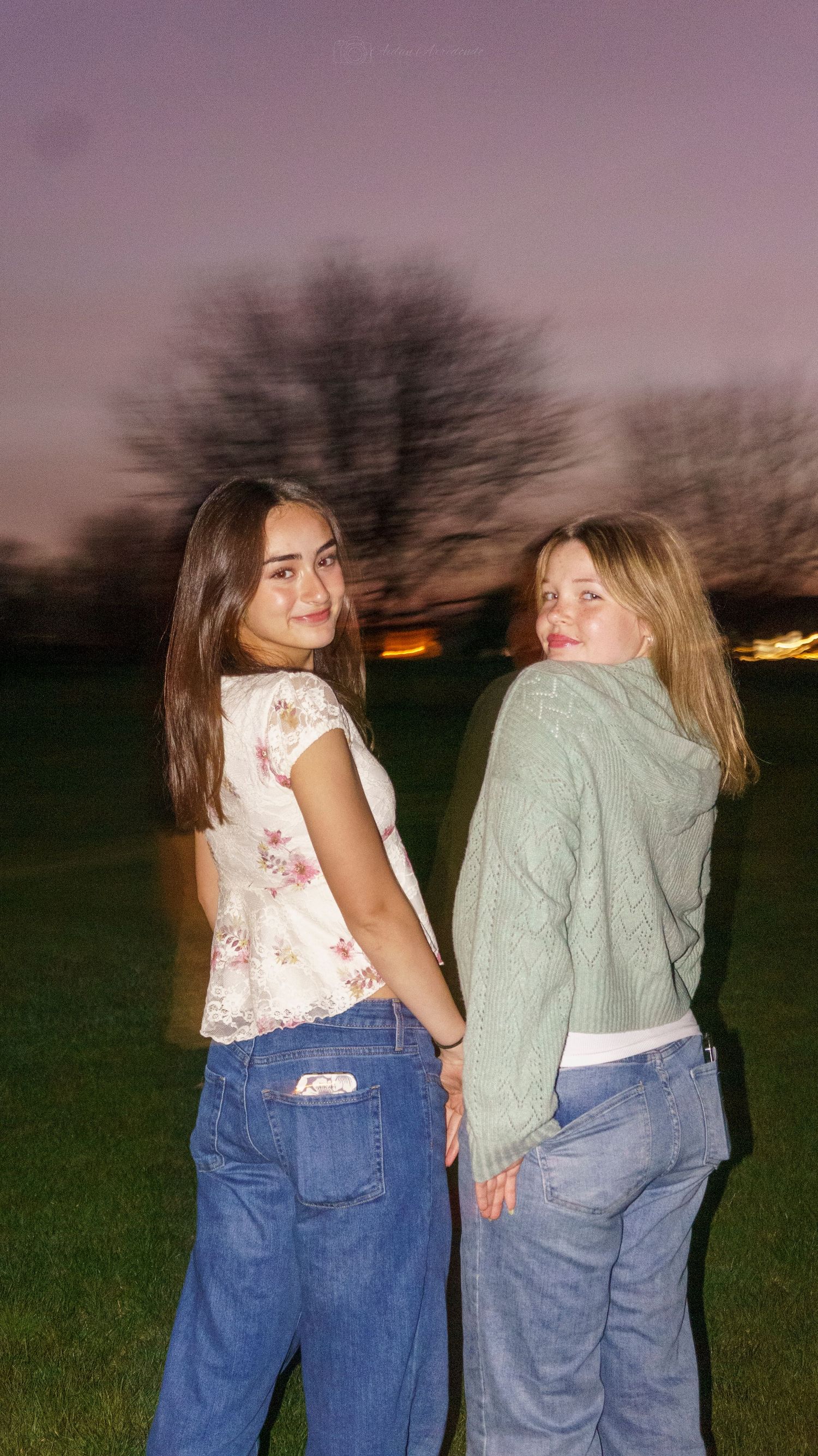 Two teenage girls in jeans posing back-to-back on grass field at dusk with purple sky.