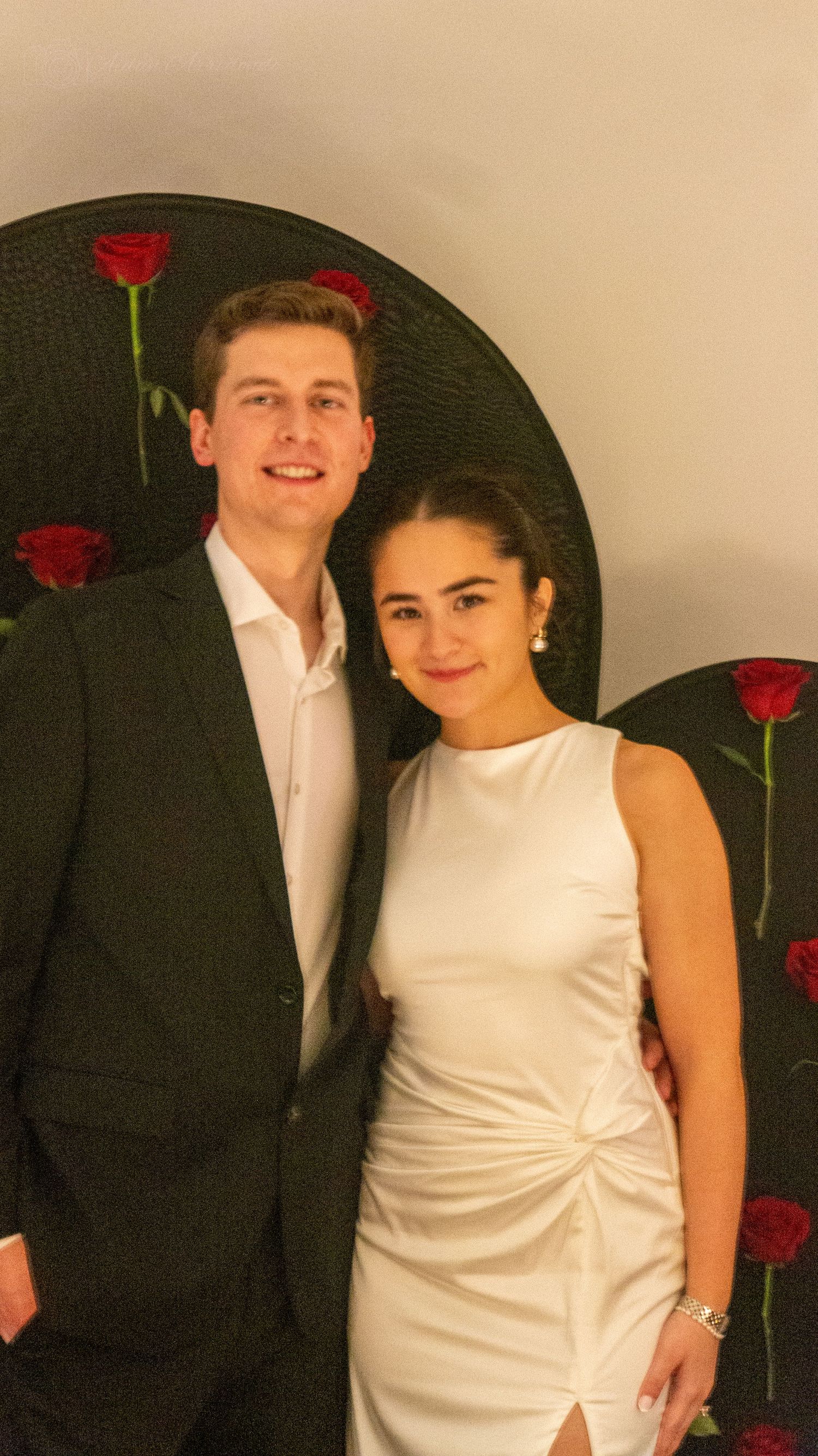 Elegant couple posing together in formal attire in front of a decorative red rose backdrop.