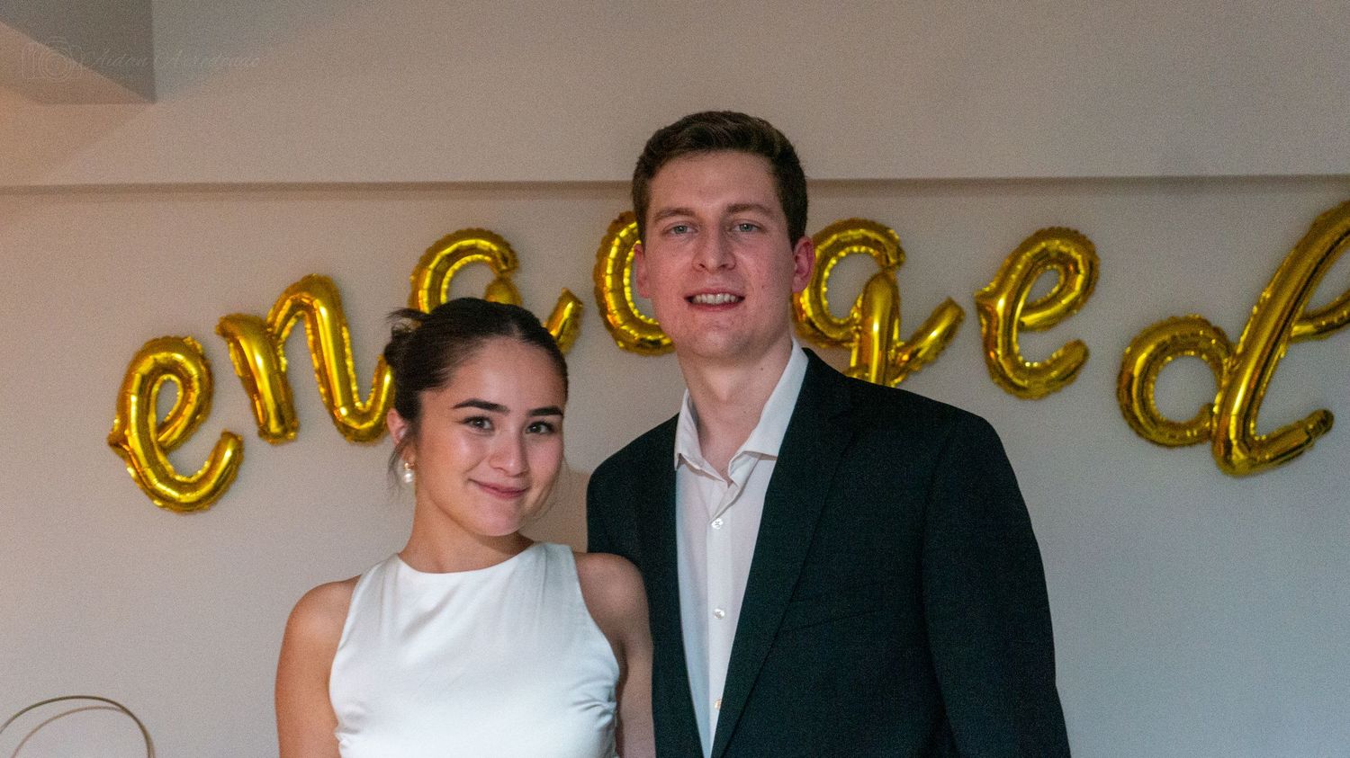 A couple poses together in front of gold engagement balloon letters at their engagement celebration.