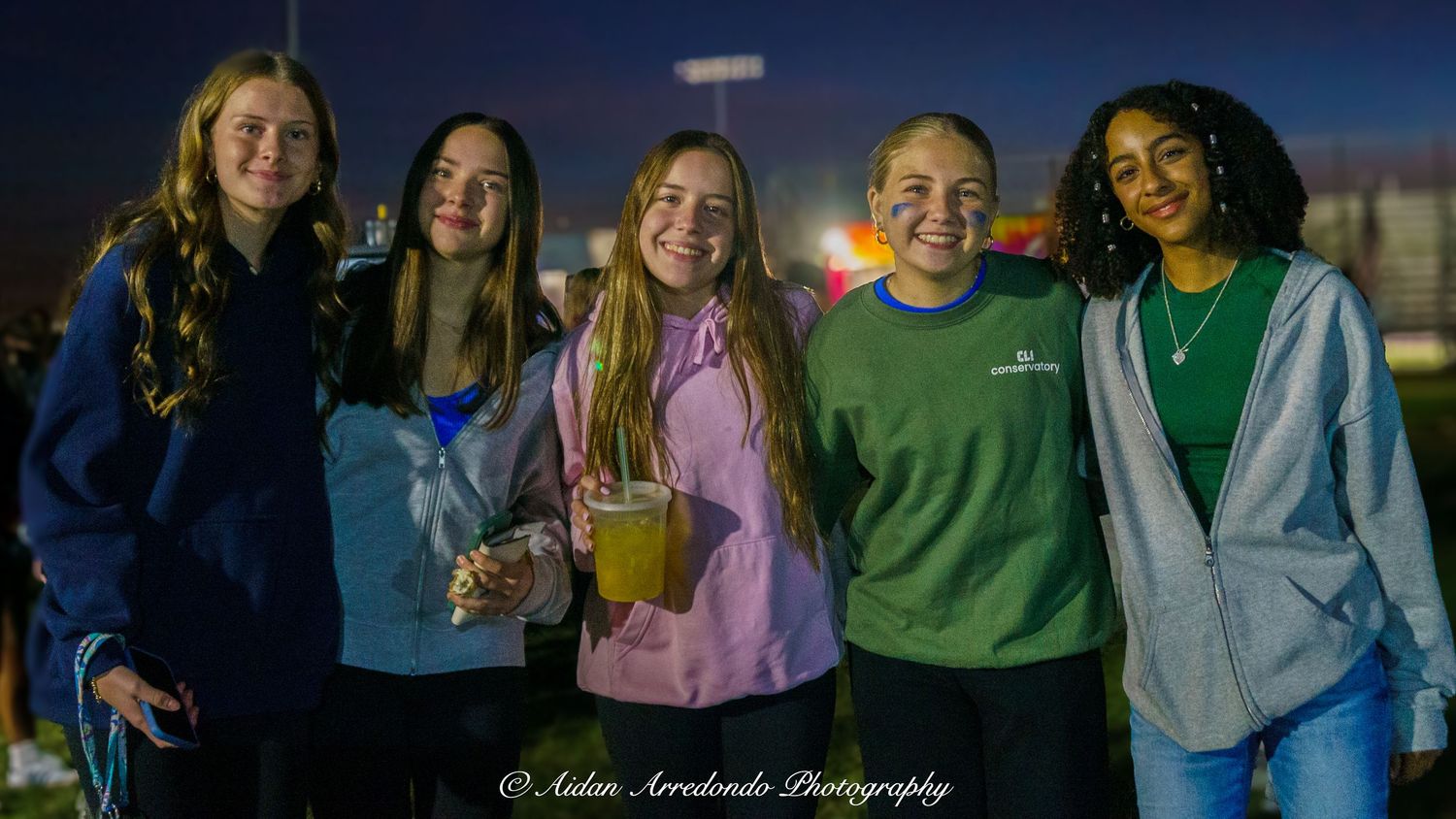Group of five smiling teenage girls posing together at an outdoor evening event or concert.