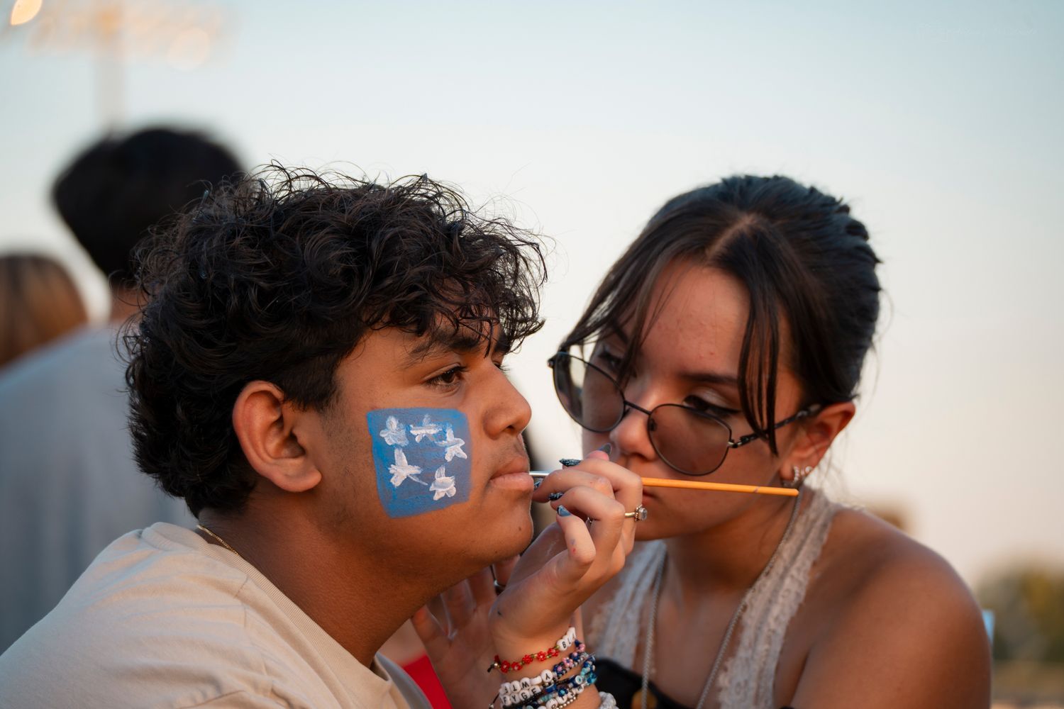 Two young people at an outdoor event as one paints stars on the others cheek.