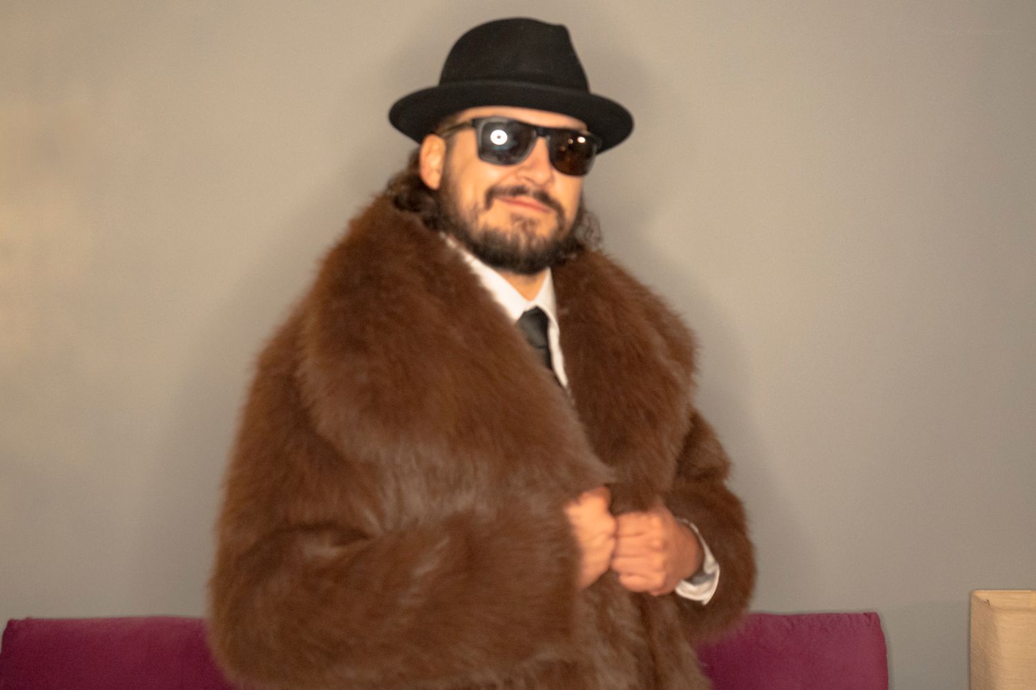 Man wearing brown fur coat, black fedora hat, and sunglasses posing confidently indoors.