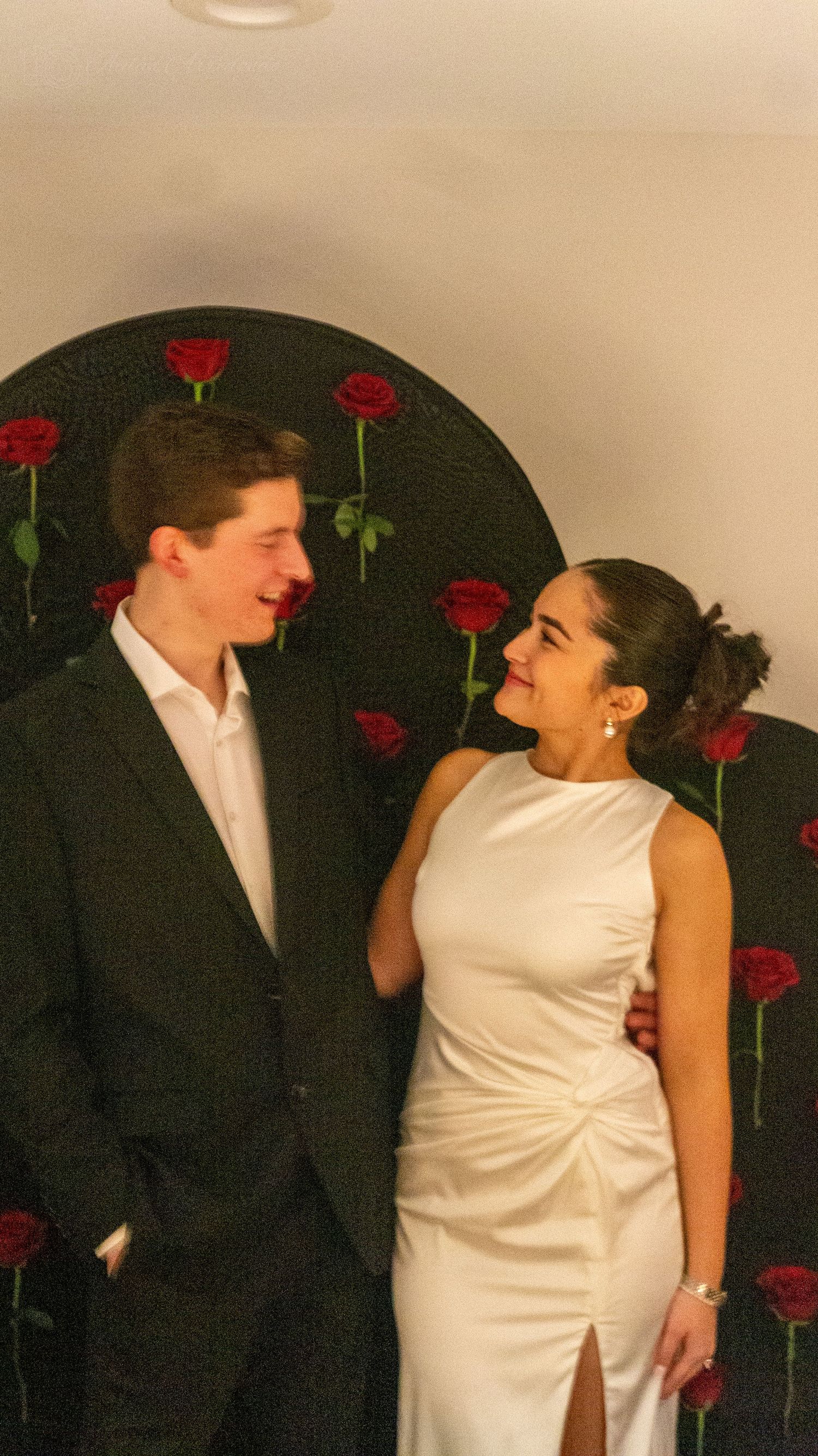 Couple smiling at each other in formal attire in front of a dark backdrop with red roses.