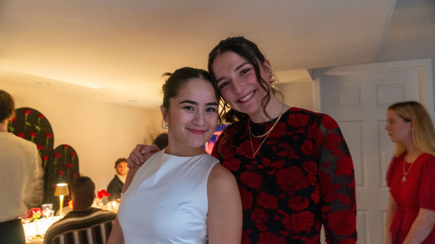 Two young women smile together at an indoor evening event with warm ambient lighting.