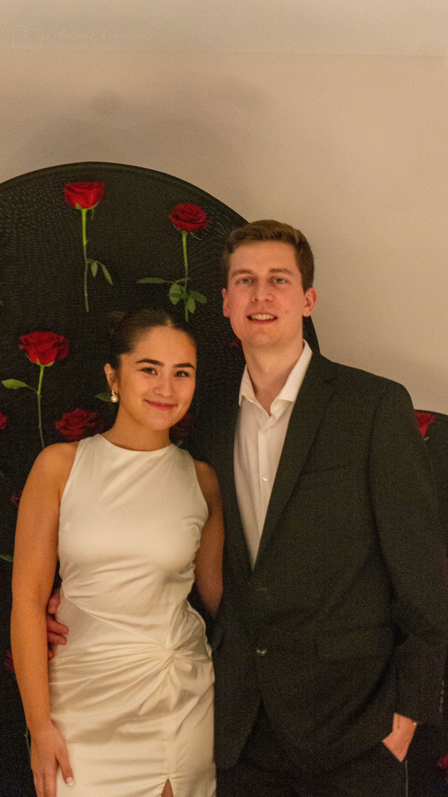 Smiling couple posing in front of red roses backdrop, woman in white dress.