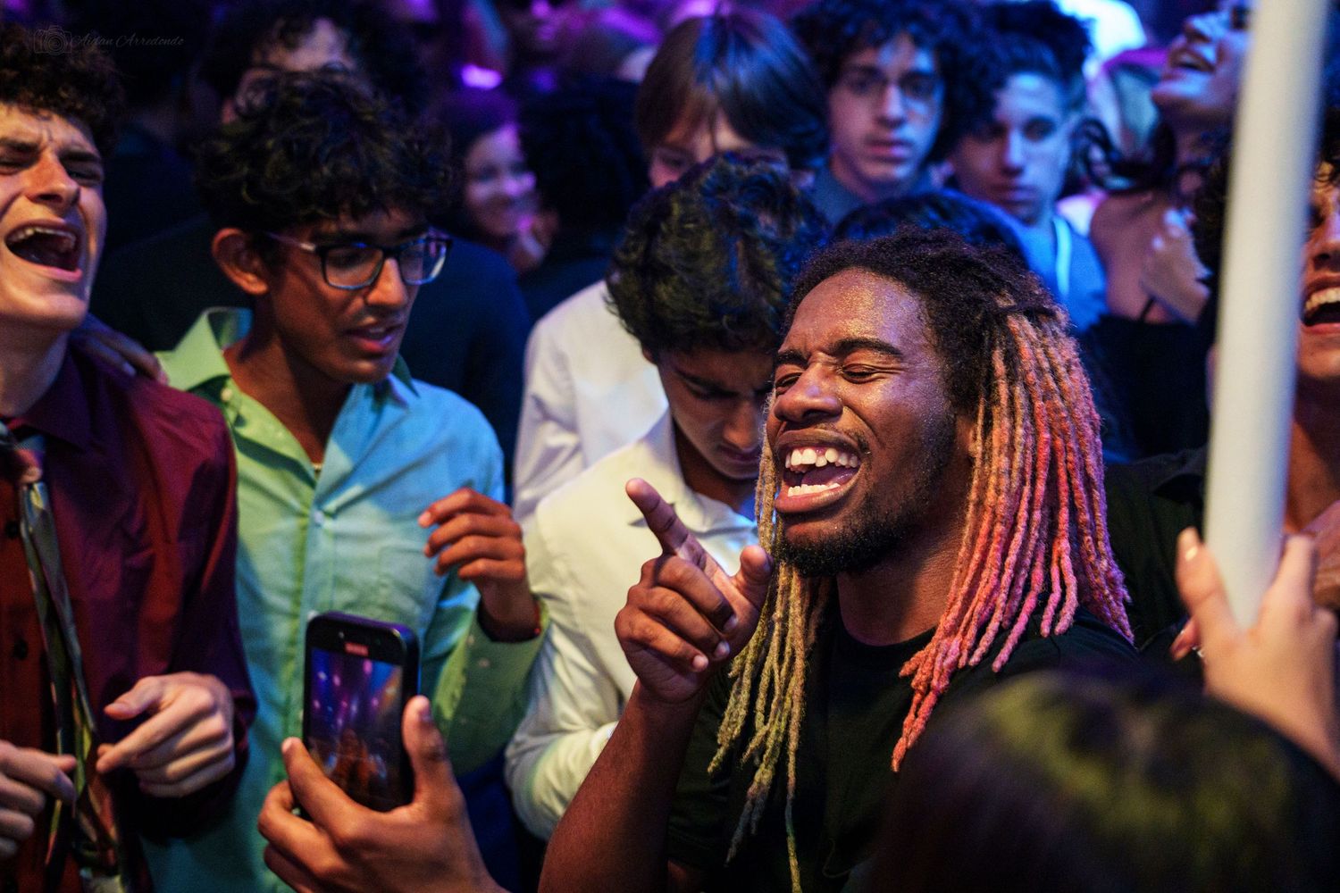 Young man with colorful dreadlocks laughing joyfully in a crowd at an indoor concert event.