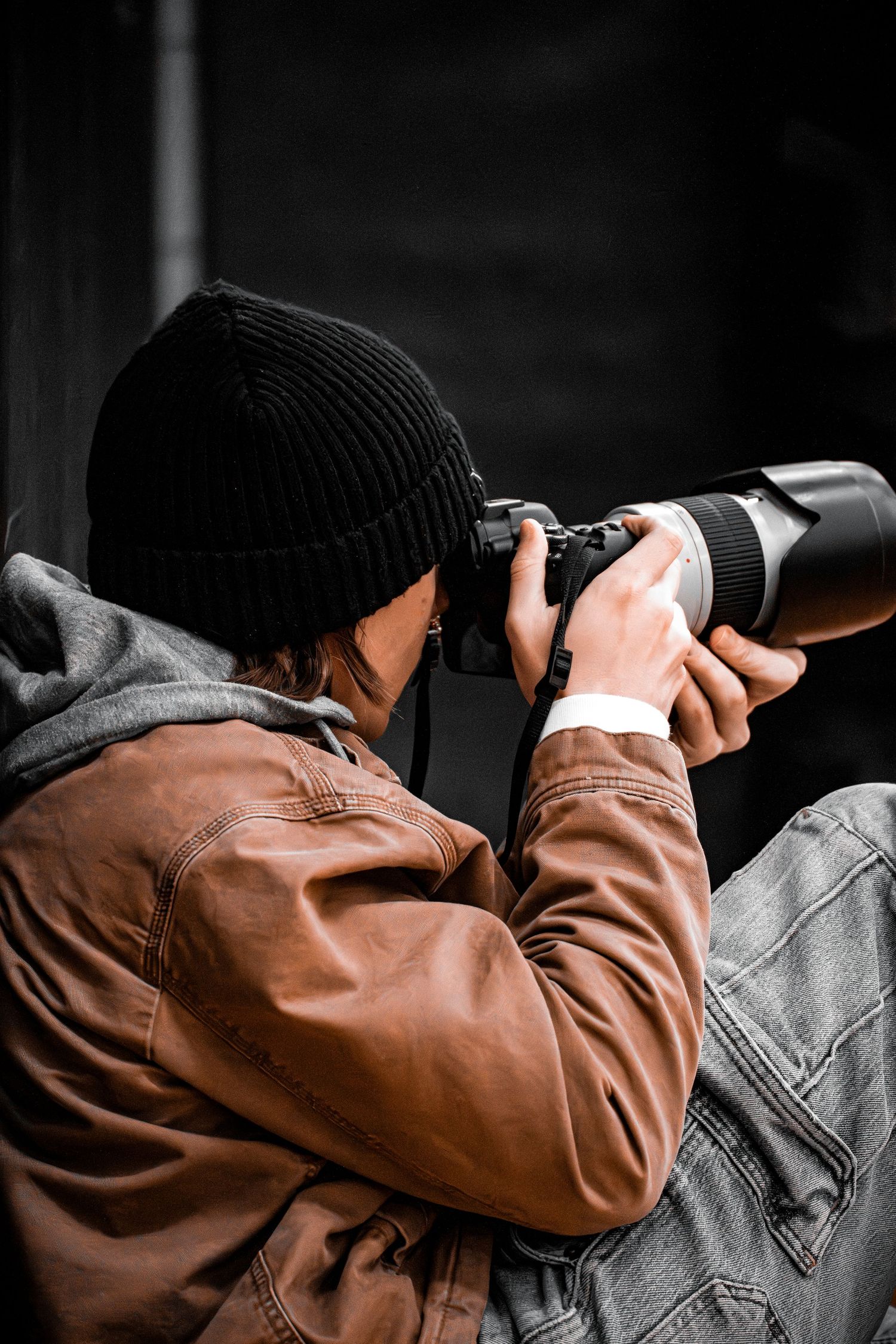 Photographer wearing brown leather jacket and black beanie aims telephoto lens camera in dramatic black and white setting.
