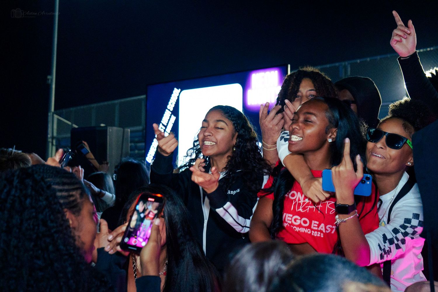 Excited crowd of young women enjoying an outdoor night concert, singing and raising hands.