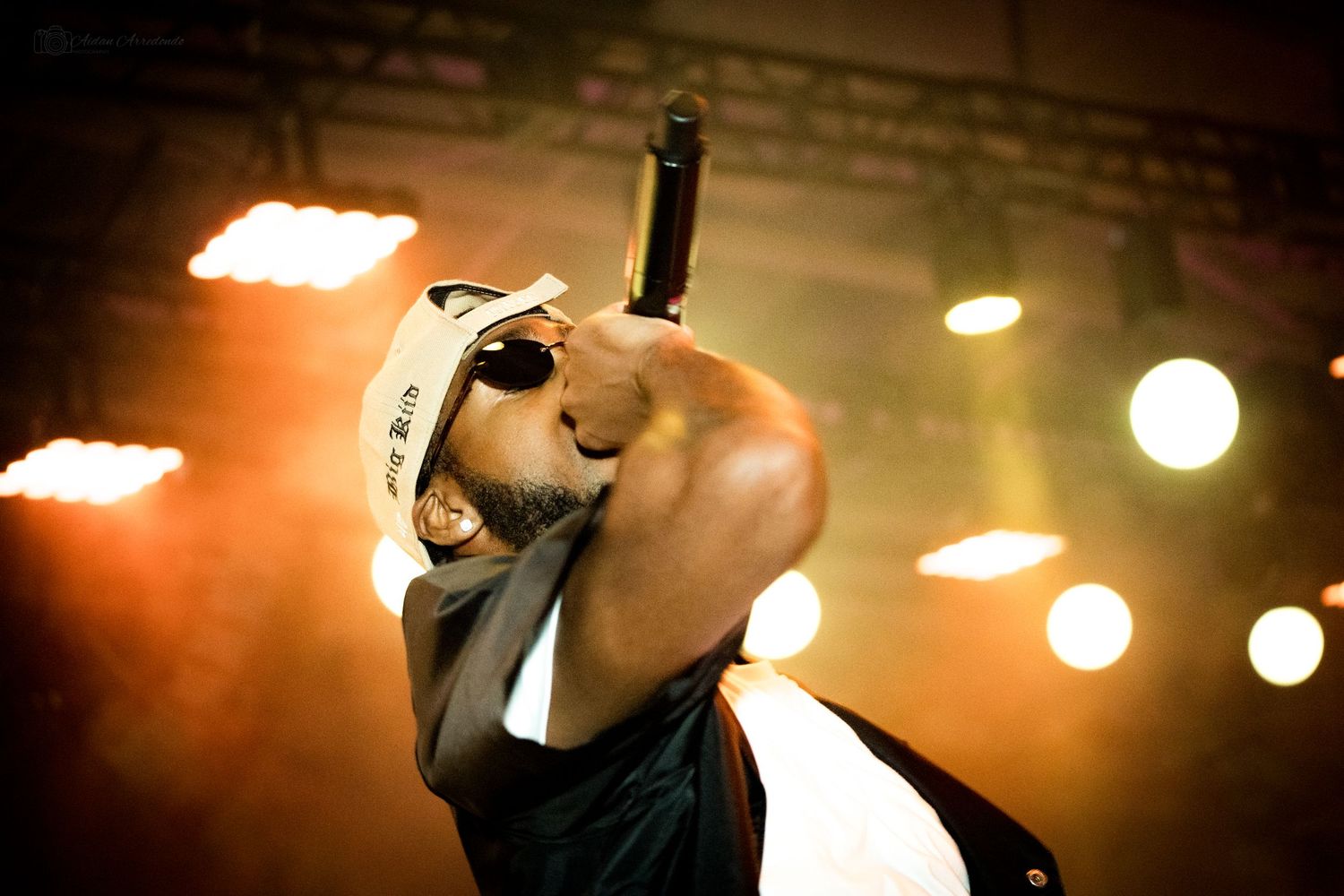 Energetic male rapper performing on stage with microphone under dramatic orange concert lighting.