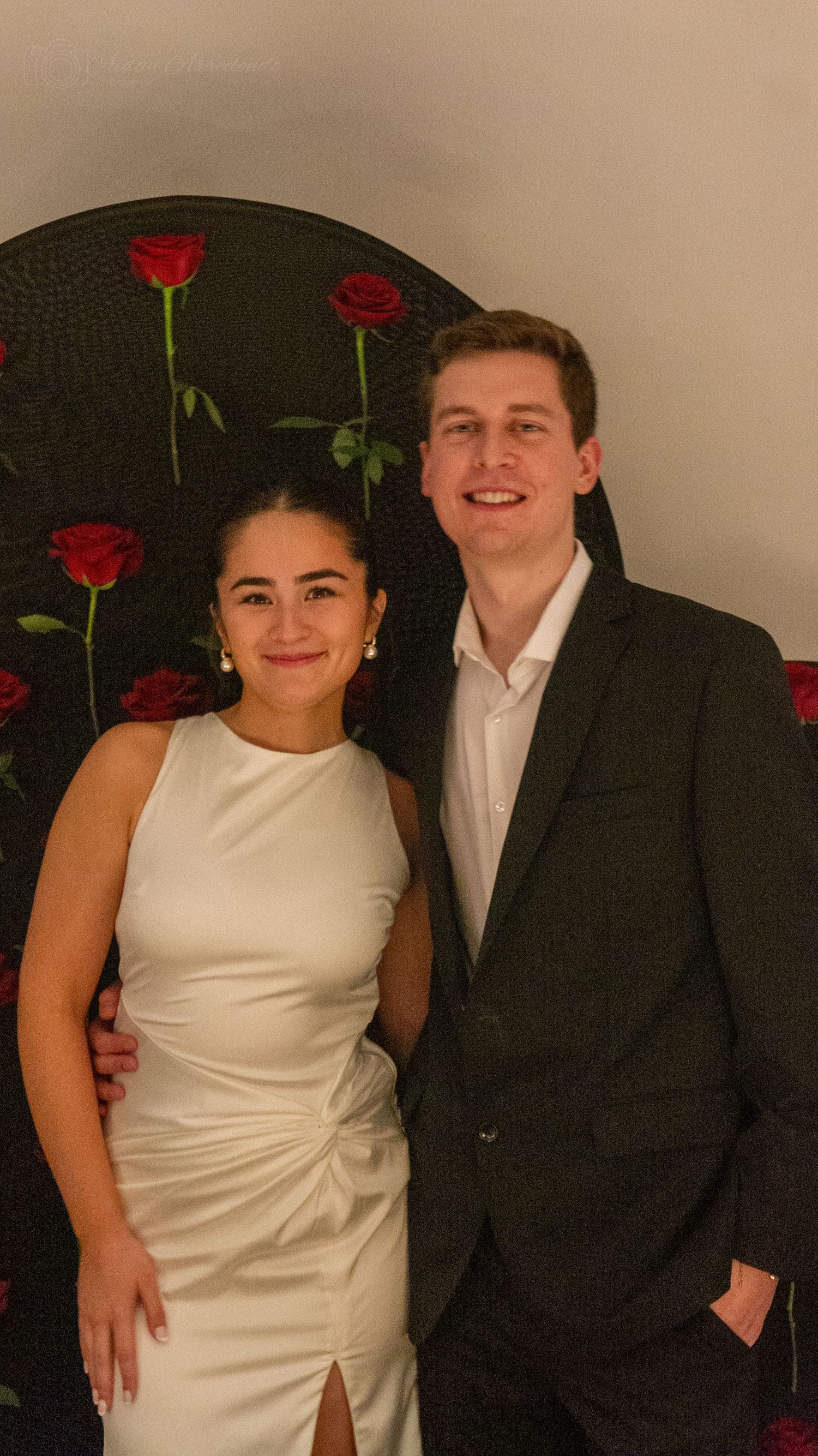 Smiling couple in formal wear posing together in front of a romantic red rose backdrop.