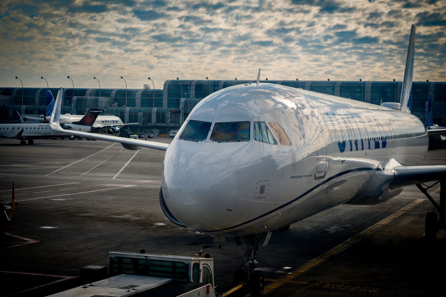 United Airlines aircraft parked at airport gate on cloudy day, with terminal and other planes visible in background.