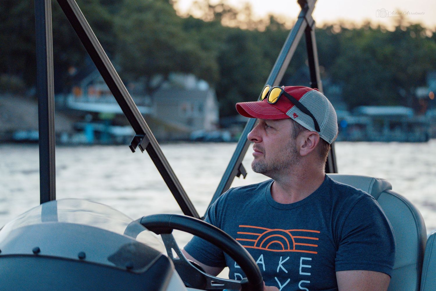 Man wearing a cap and t-shirt drives a pontoon boat on a lake at sunset.