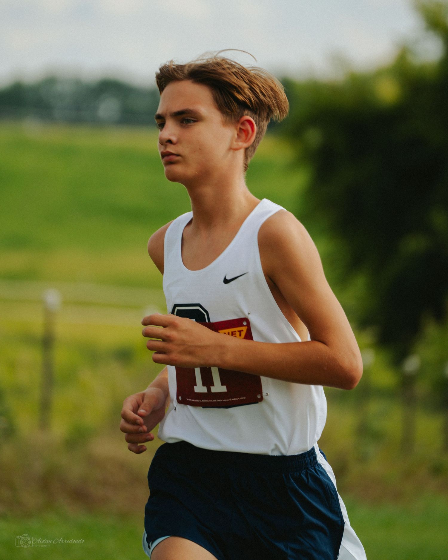 Young male cross country runner in white Nike jersey number 11 races through a green outdoor course.