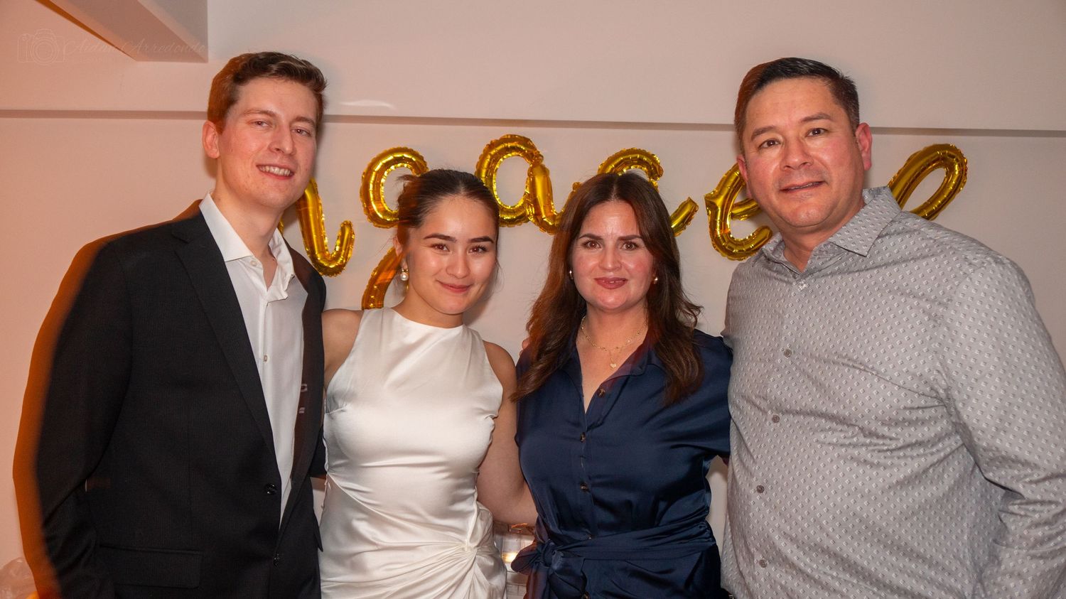 Two couples smile together at an engagement party with gold letter balloons in the background.