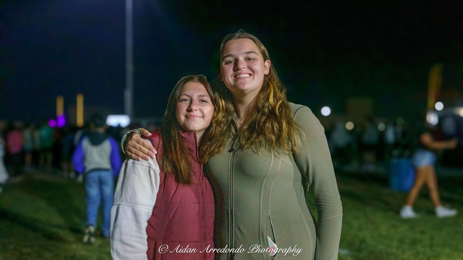 Two smiling young women posing together at an outdoor nighttime event on a green field.