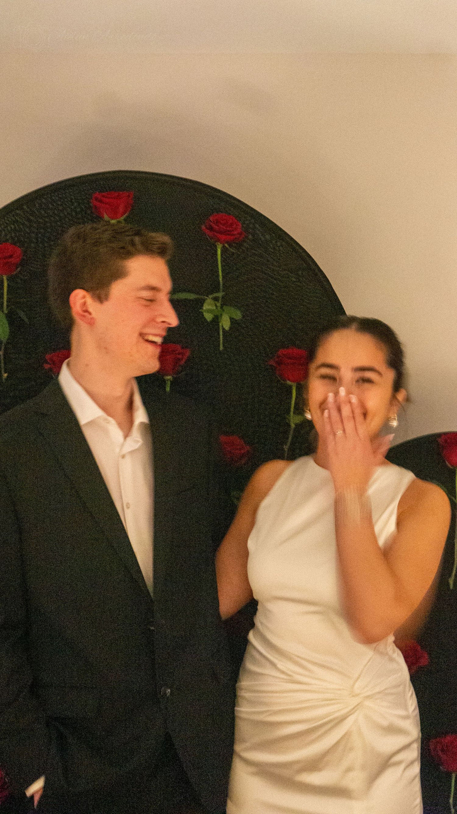 Laughing engaged couple standing in front of a decorative red rose backdrop at their engagement party.