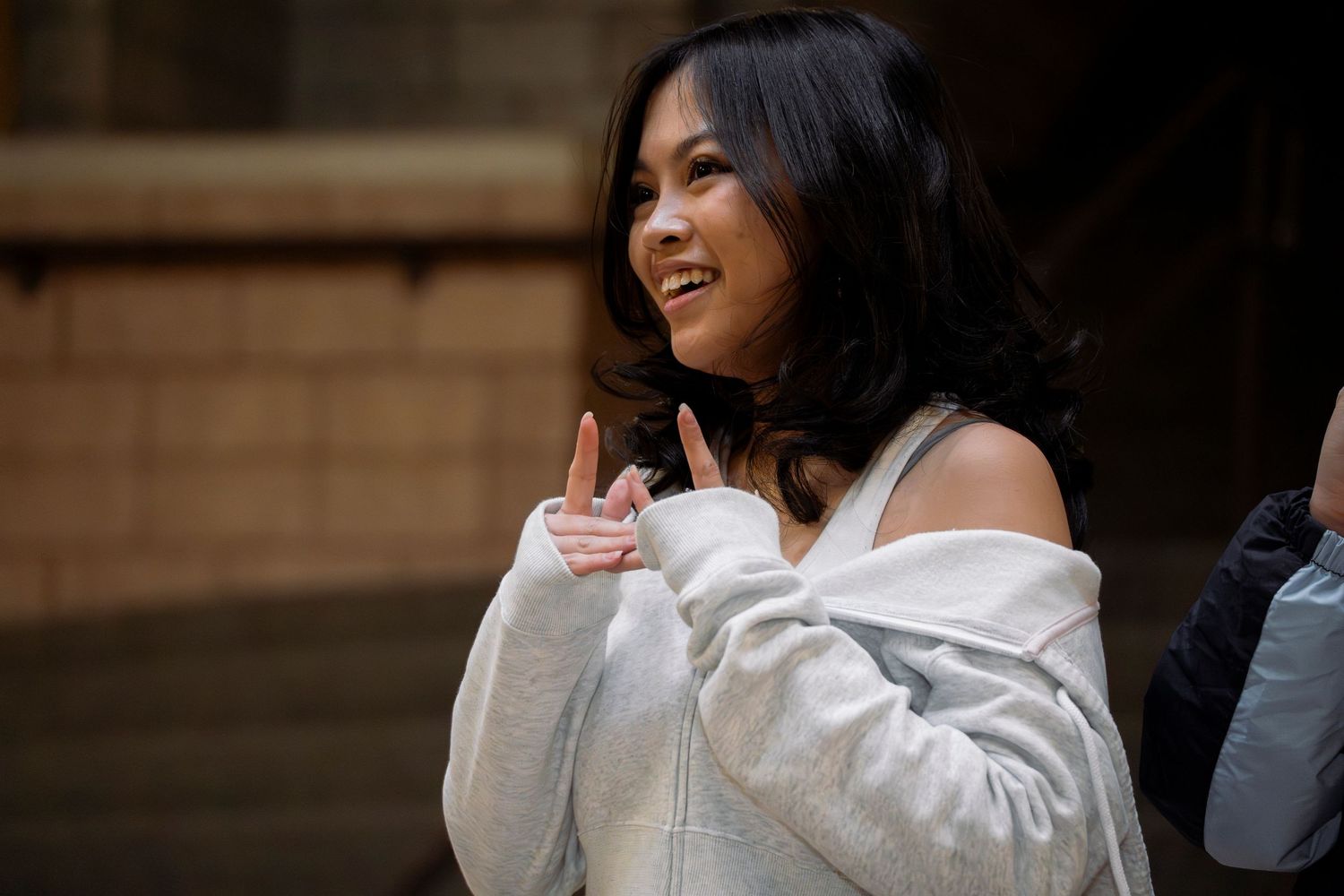 Young Asian woman in white off-shoulder hoodie smiling and gesturing outdoors.
