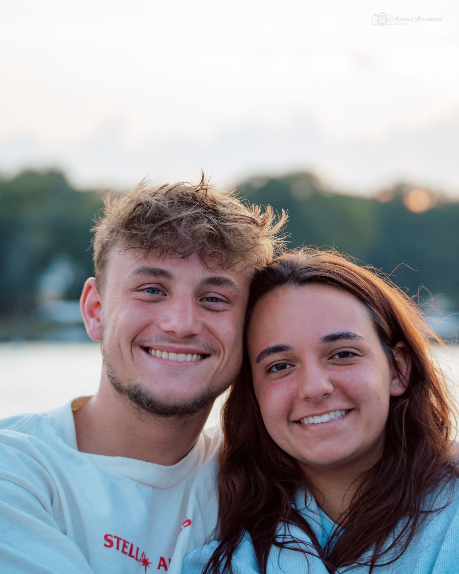 A smiling young couple poses closely together outdoors near a lake at sunset.
