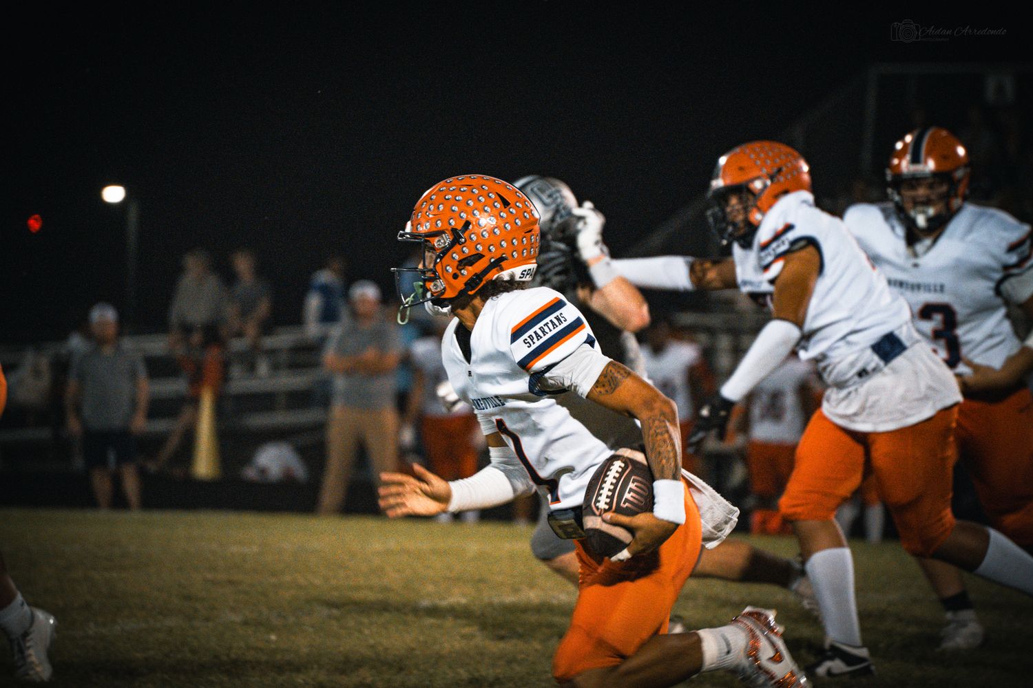 High school football player in orange and white uniform runs with the ball during a night game.