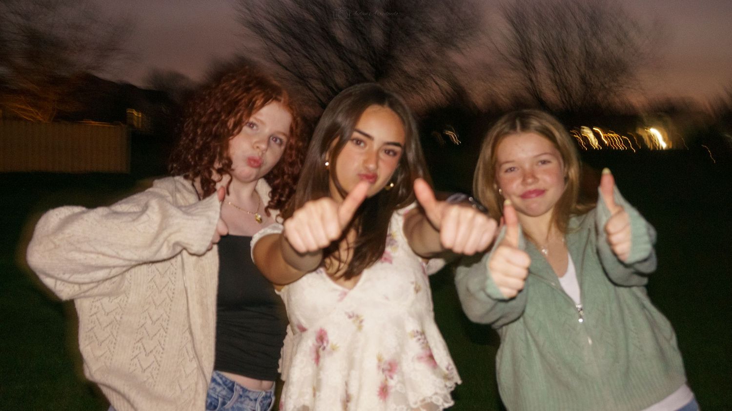 Three teenage girls giving thumbs up and smiling together outdoors at dusk on green lawn.