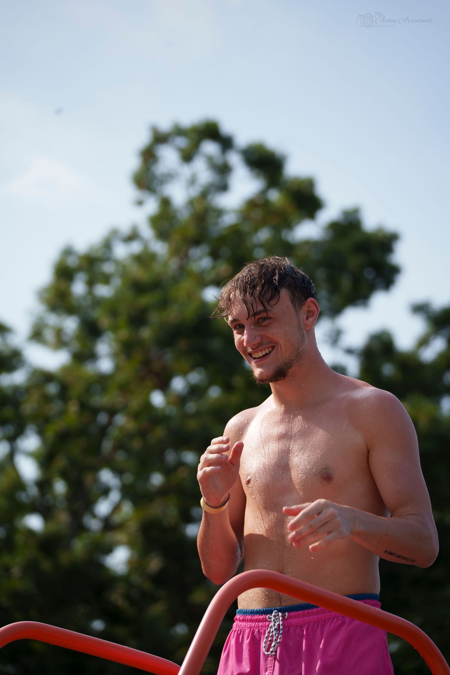 Shirtless smiling young man with wet hair standing outdoors near red railing on sunny day.