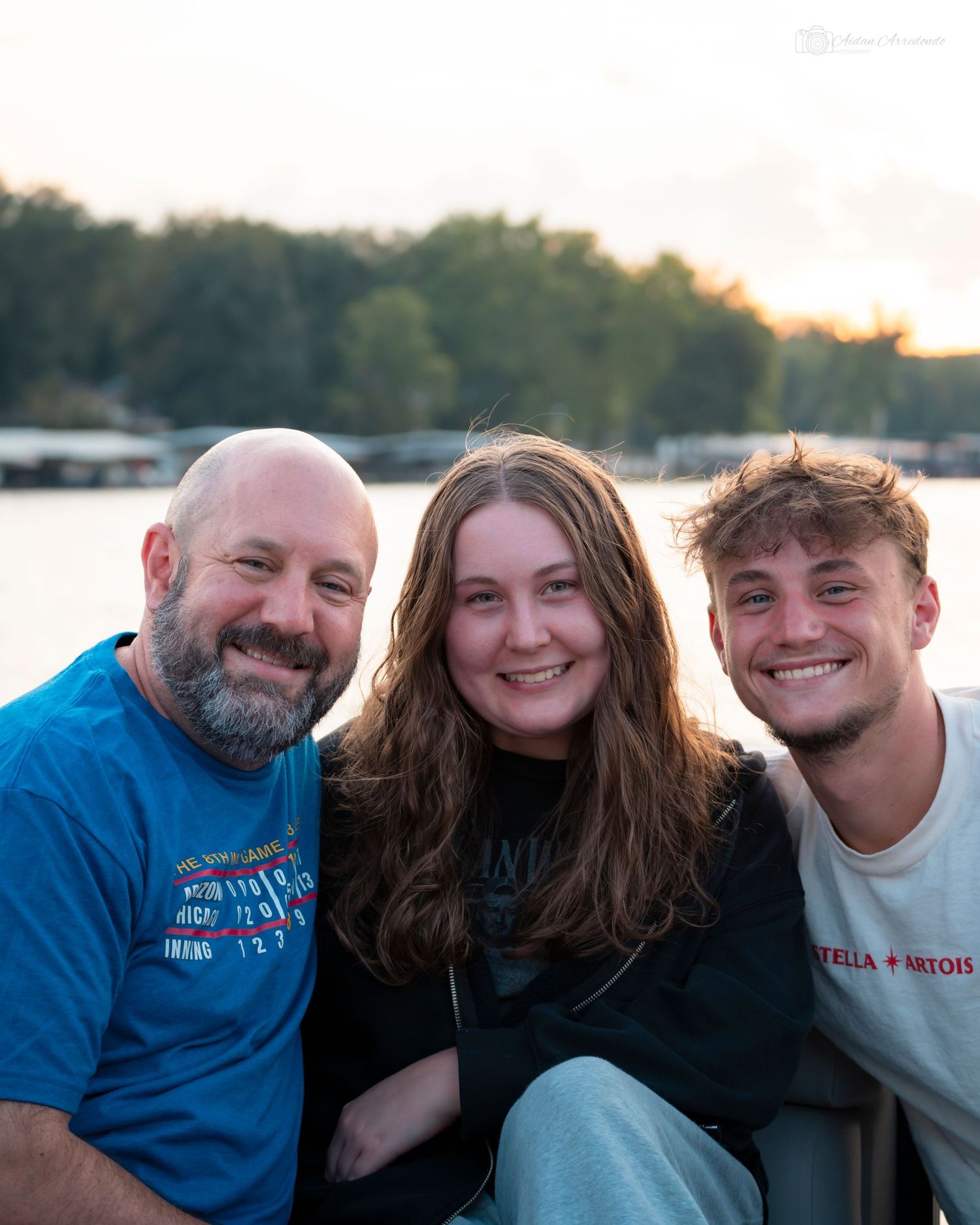 Family of three smiling together at sunset near a lake, father in blue shirt with two young adults.