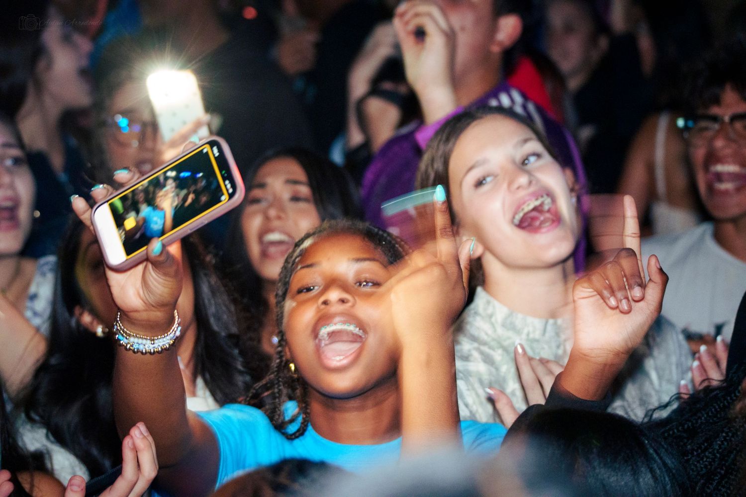 Excited young fans in a concert crowd, one taking a selfie with a glowing phone screen.