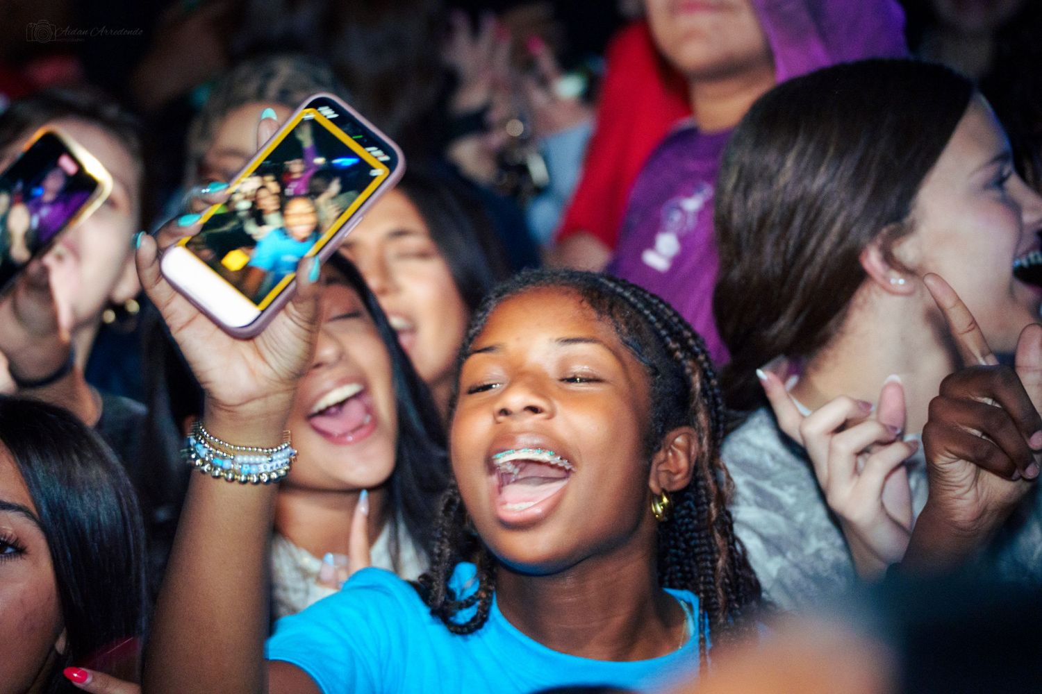 Excited young girl with braces smiles at concert while crowd holds up smartphones to record.