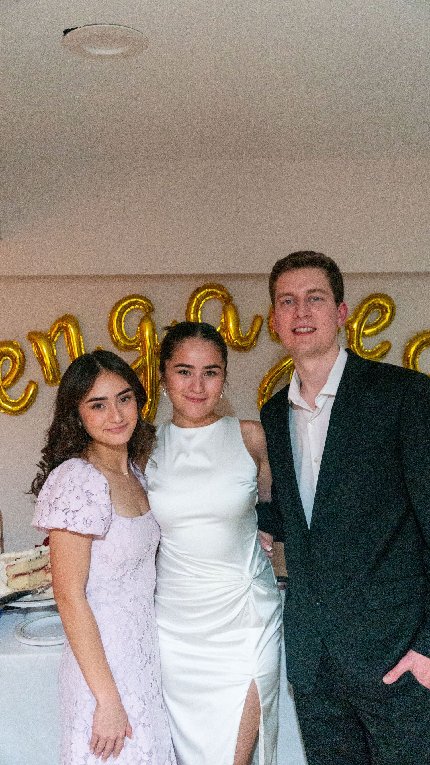 Three people pose at an engagement party with gold balloon letters spelling engaged on the wall behind them.