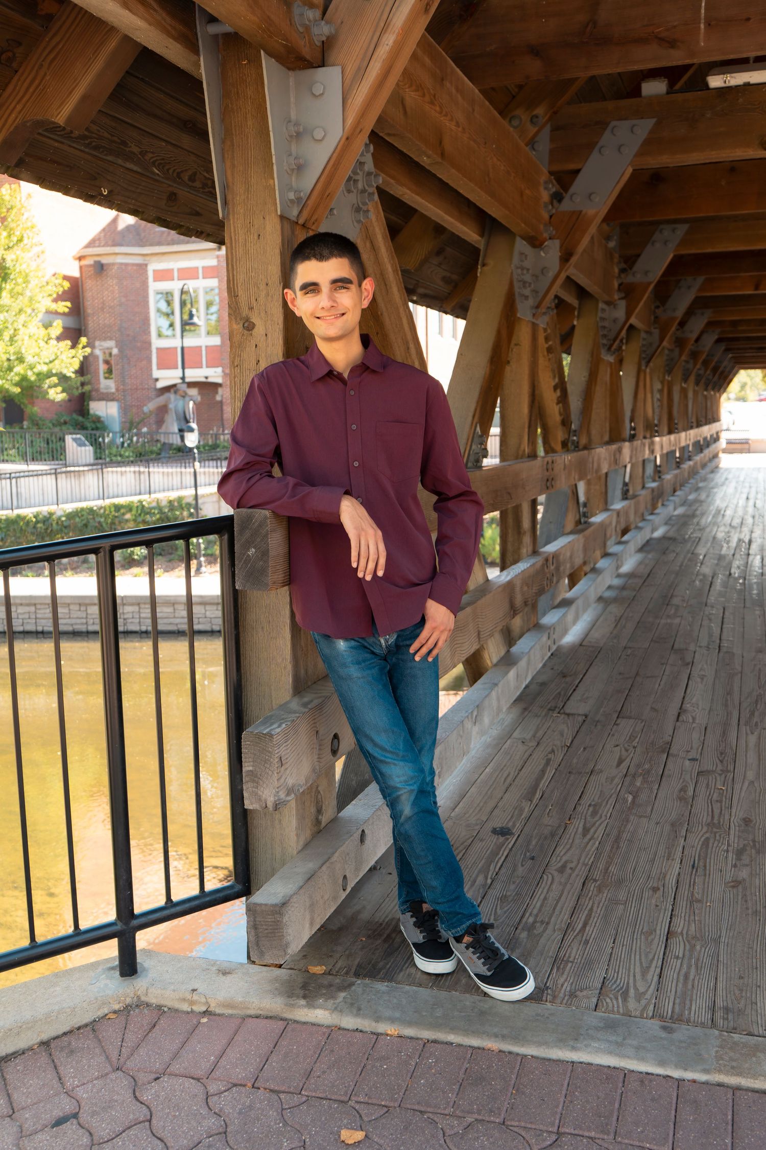 Teen boy in burgundy shirt and jeans leaning on railing of wooden covered bridge smiling.