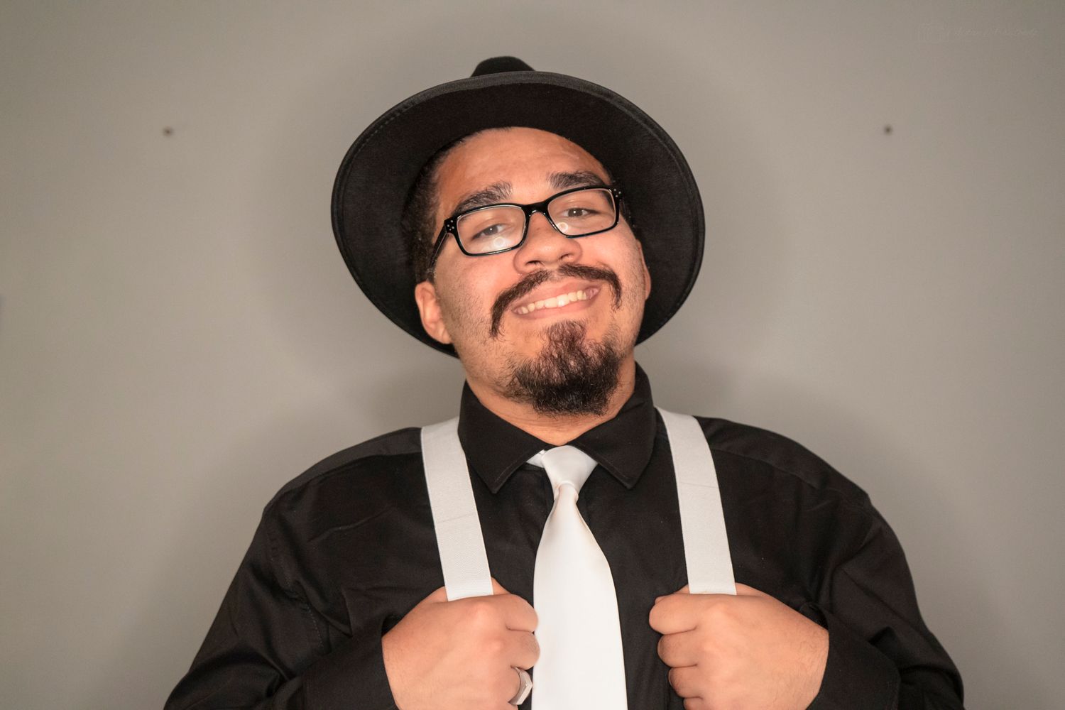 Stylish man in black hat, glasses, and suspenders smiling against gray studio background.