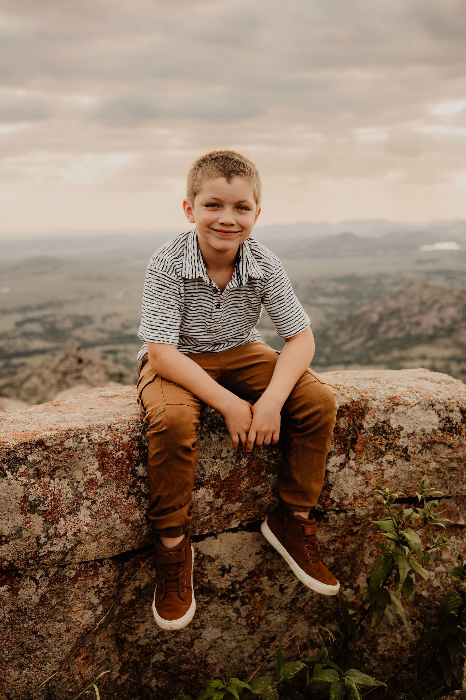 Young person in striped polo sits on rocky overlook with scenic mountain vista behind.