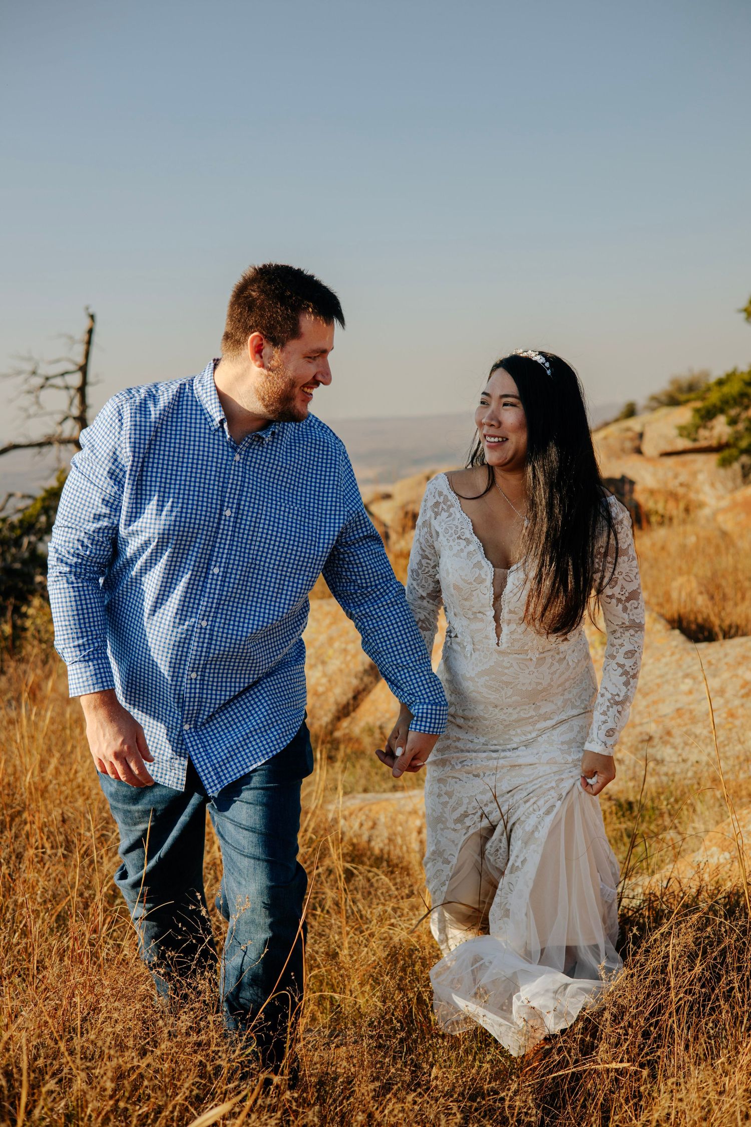 A couple walks hand in hand through golden grass with mountain views in the background.