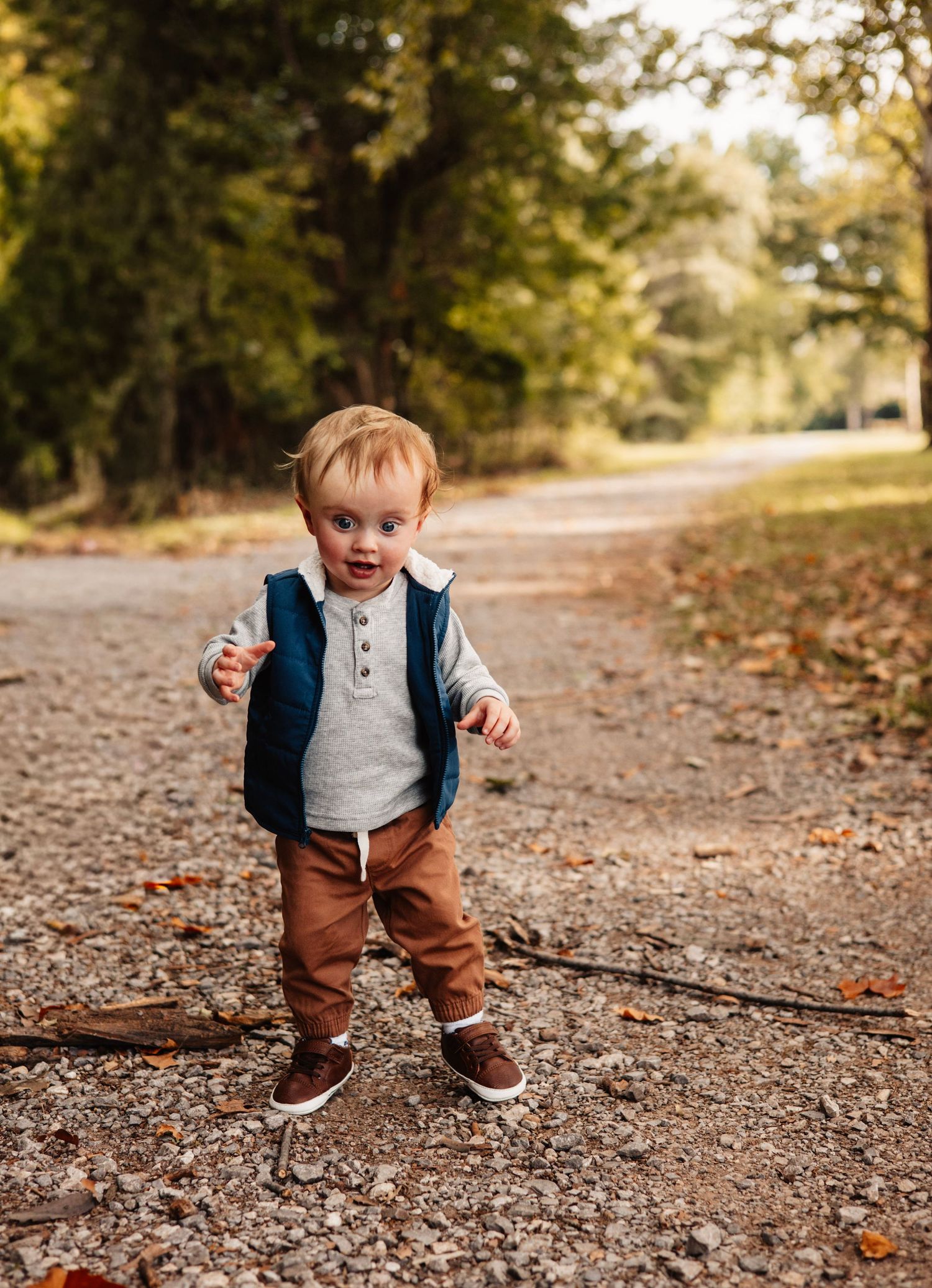 Toddler in vest and brown pants stands confidently on autumn path surrounded by fallen leaves.