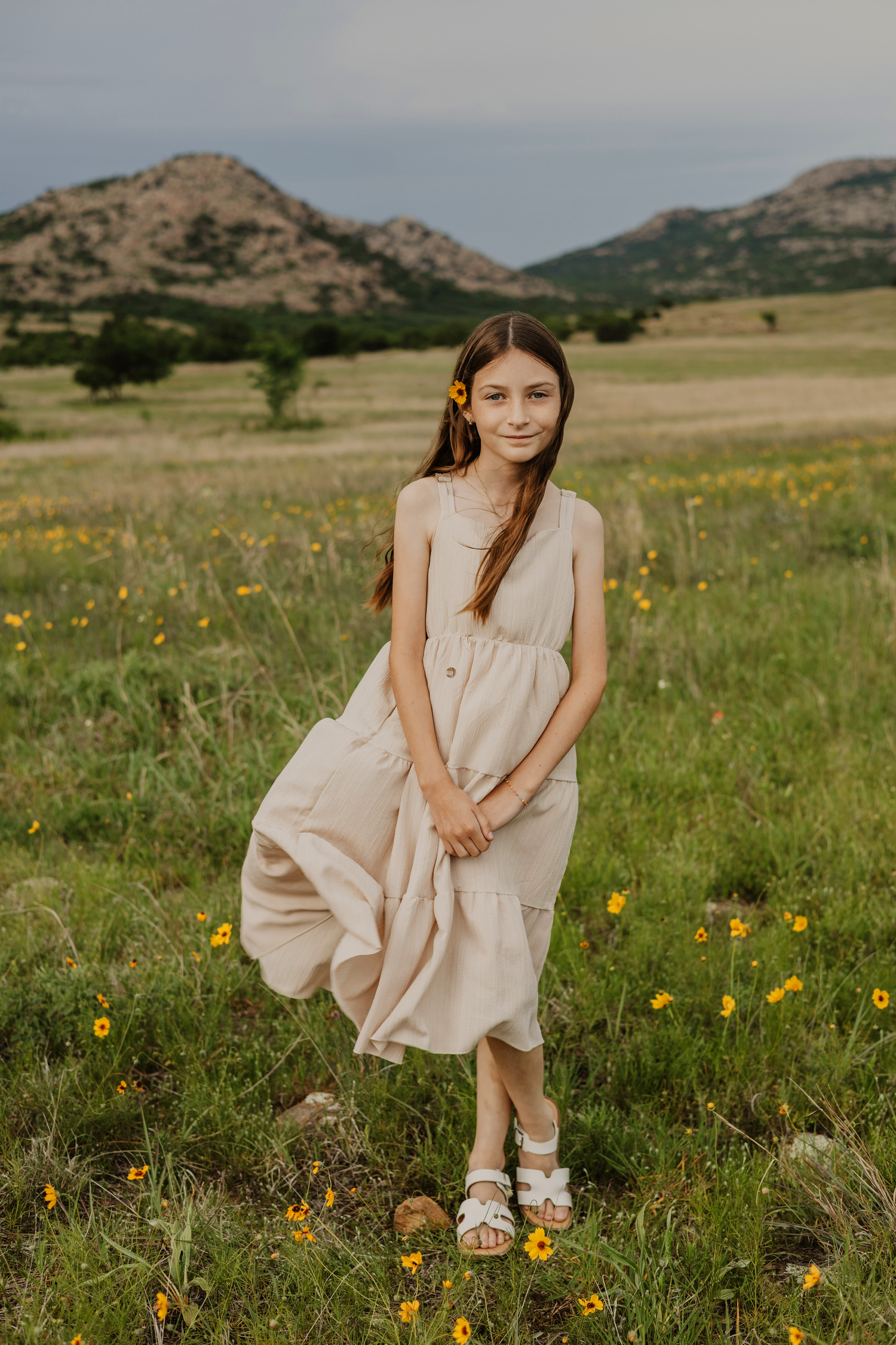 A young girl in a beige dress with a yellow flower in her hair stands in a wildflower meadow with mountains behind her.