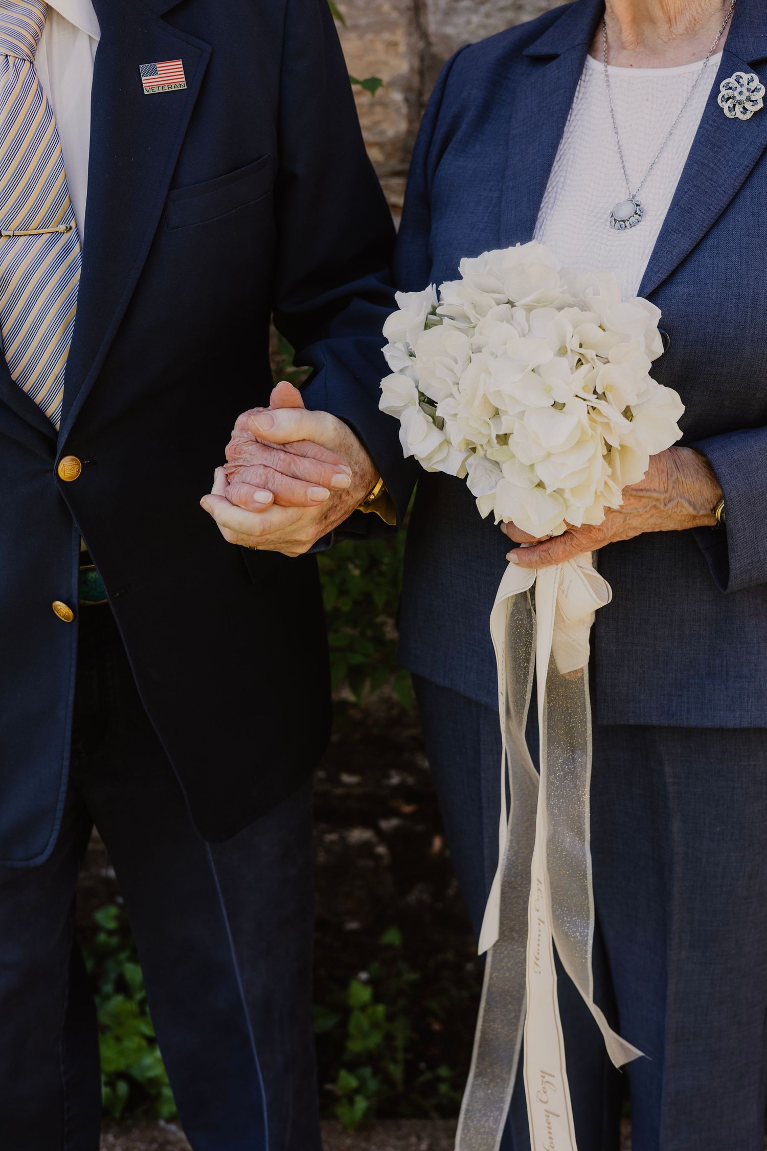 Person in navy suit holds elegant white floral bouquet with ribbon details.