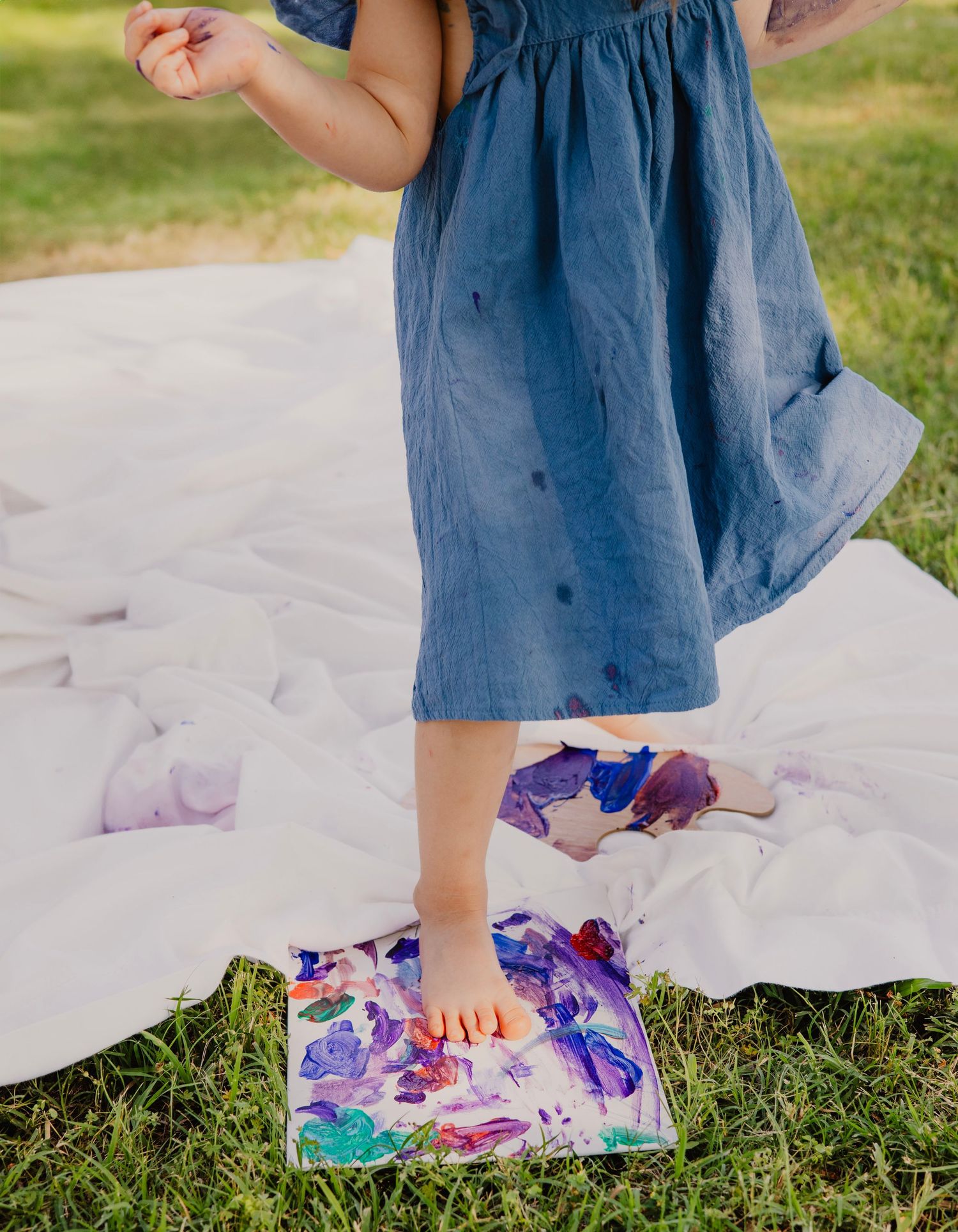 Small feet stand on colorful purple and pink watercolor artwork while wearing a flowing blue dress on a white blanket outdoors.
