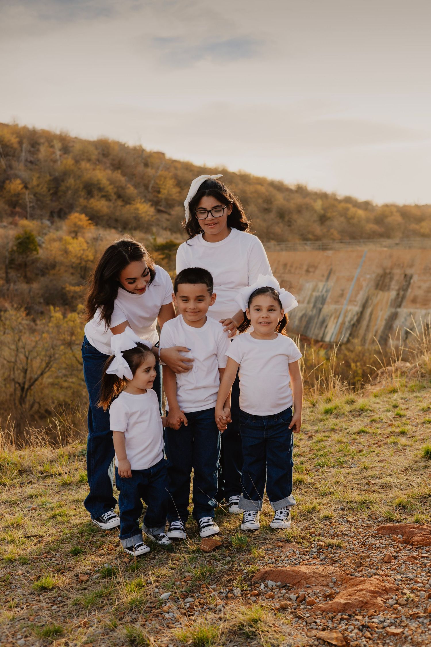 Five children in matching white shirts and jeans pose together outdoors on a hill with autumn foliage in the background.