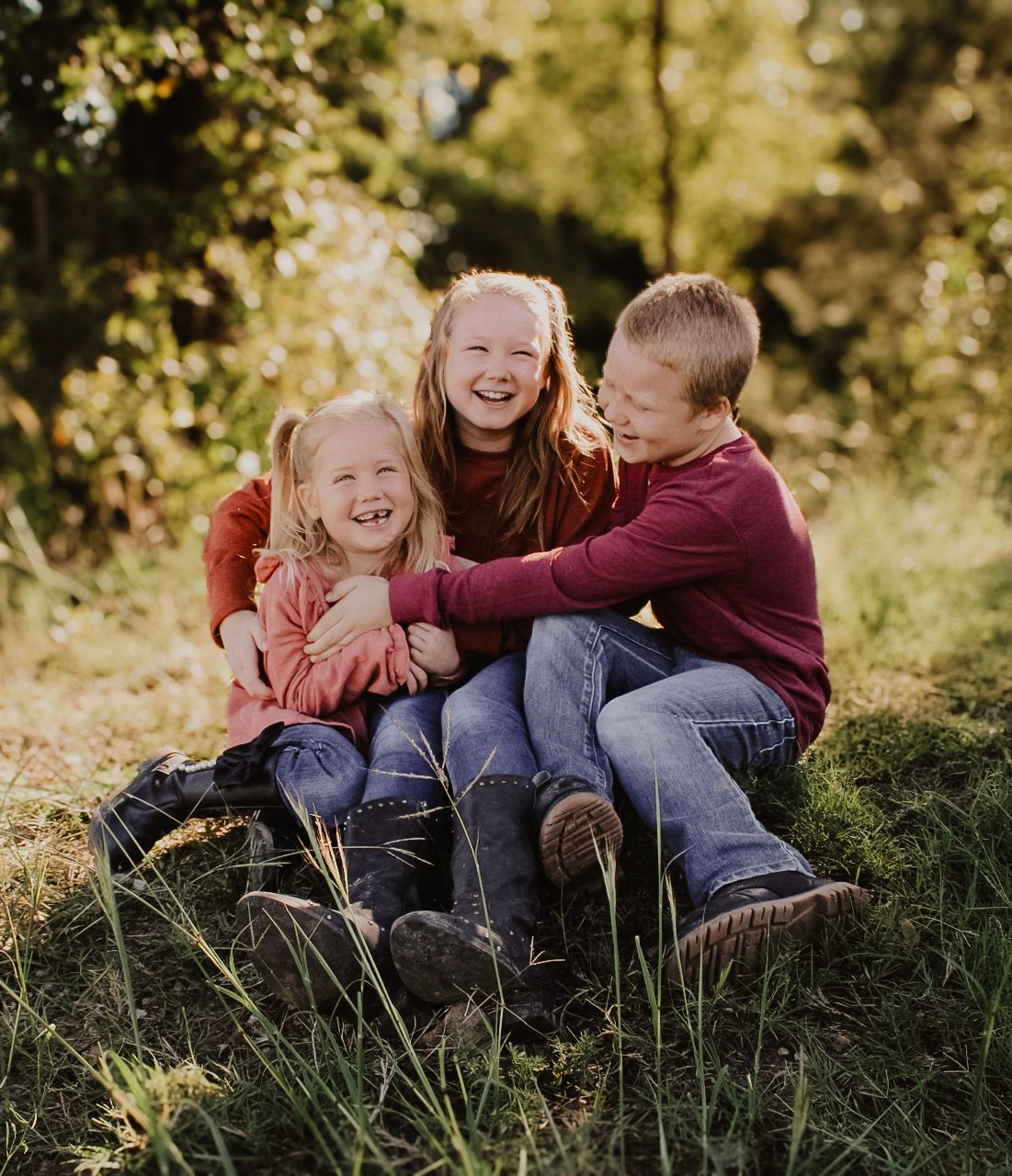 Three children laughing and hugging while sitting together on grass in autumn sunlight.