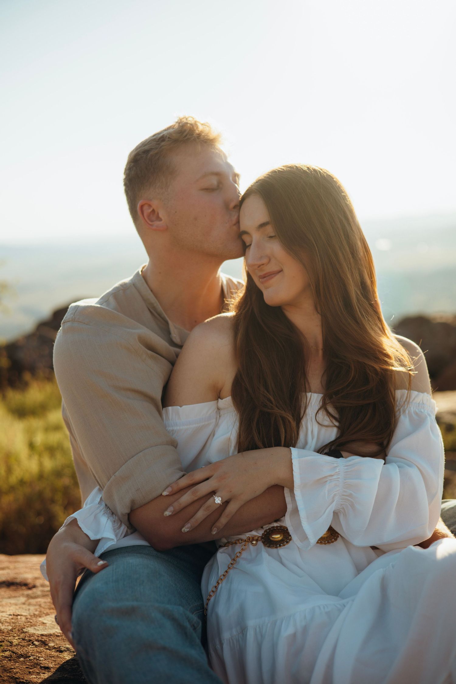 A gentle embrace between a couple in white and beige attire on a mountain overlook.